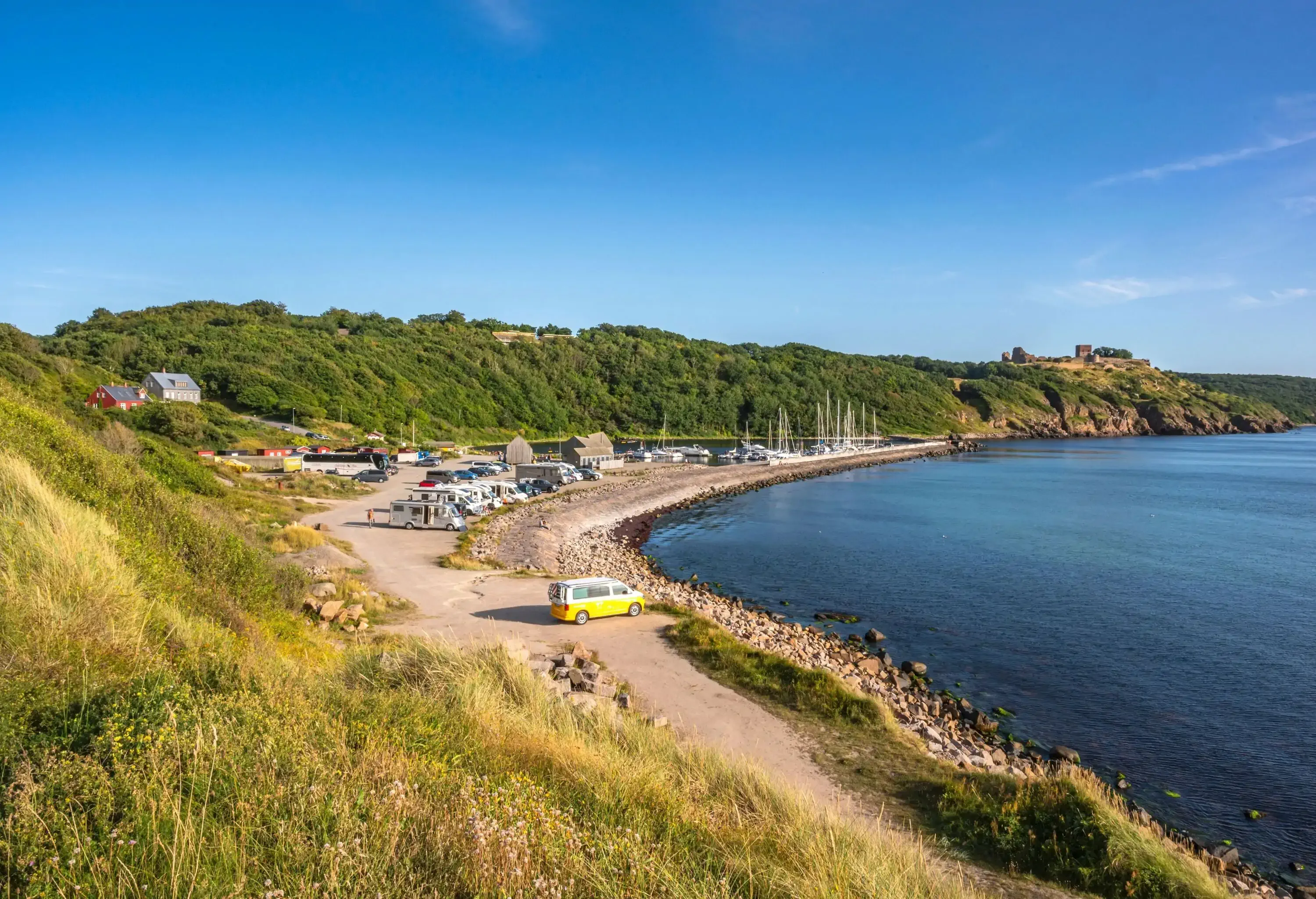 a marina with boats docked along a curved shoreline, surrounded by green hills and a parking area with camper vans.