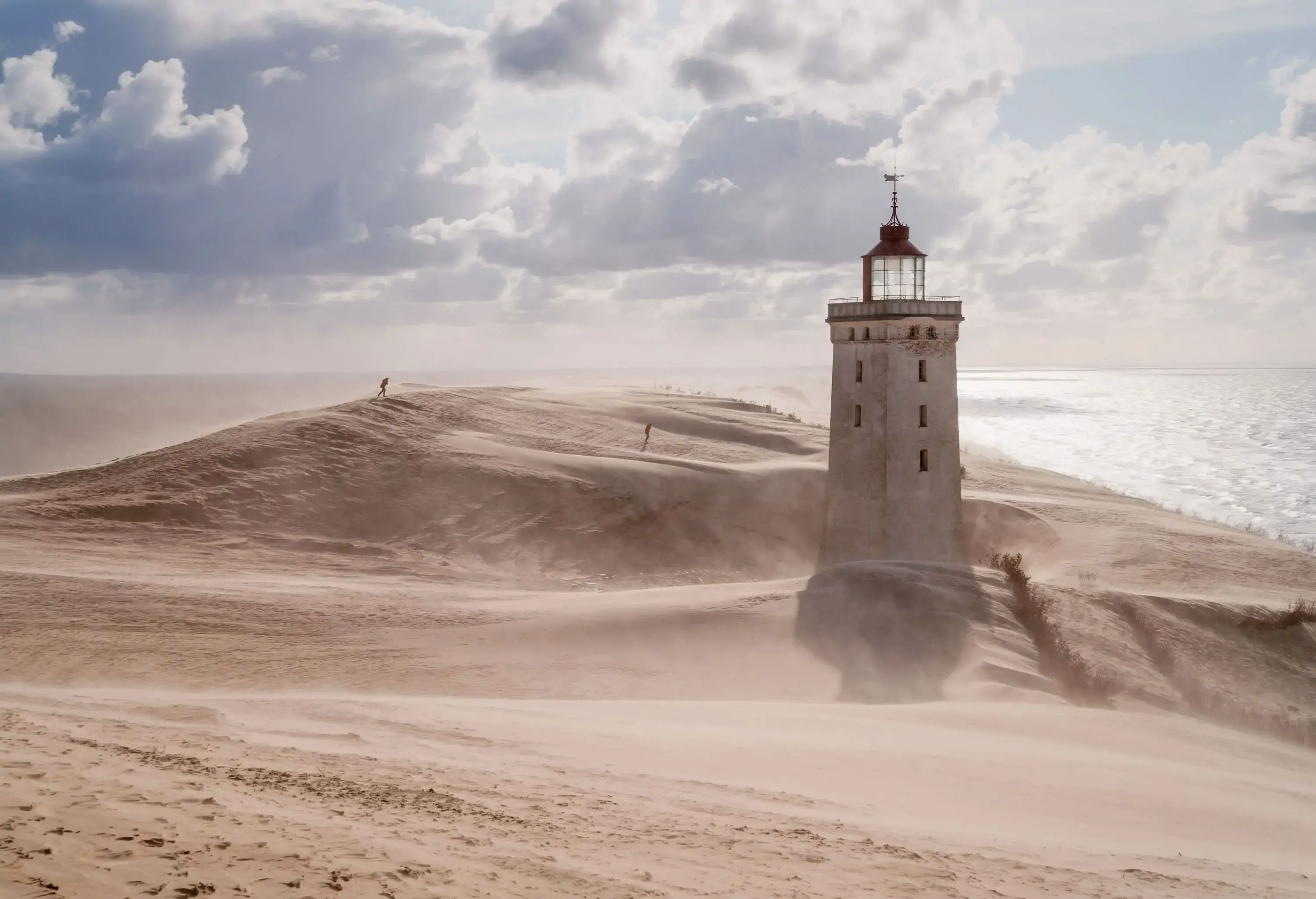 A sunlit square lighthouse with a balcony and a lantern on a rugged dune overlooking the sea.