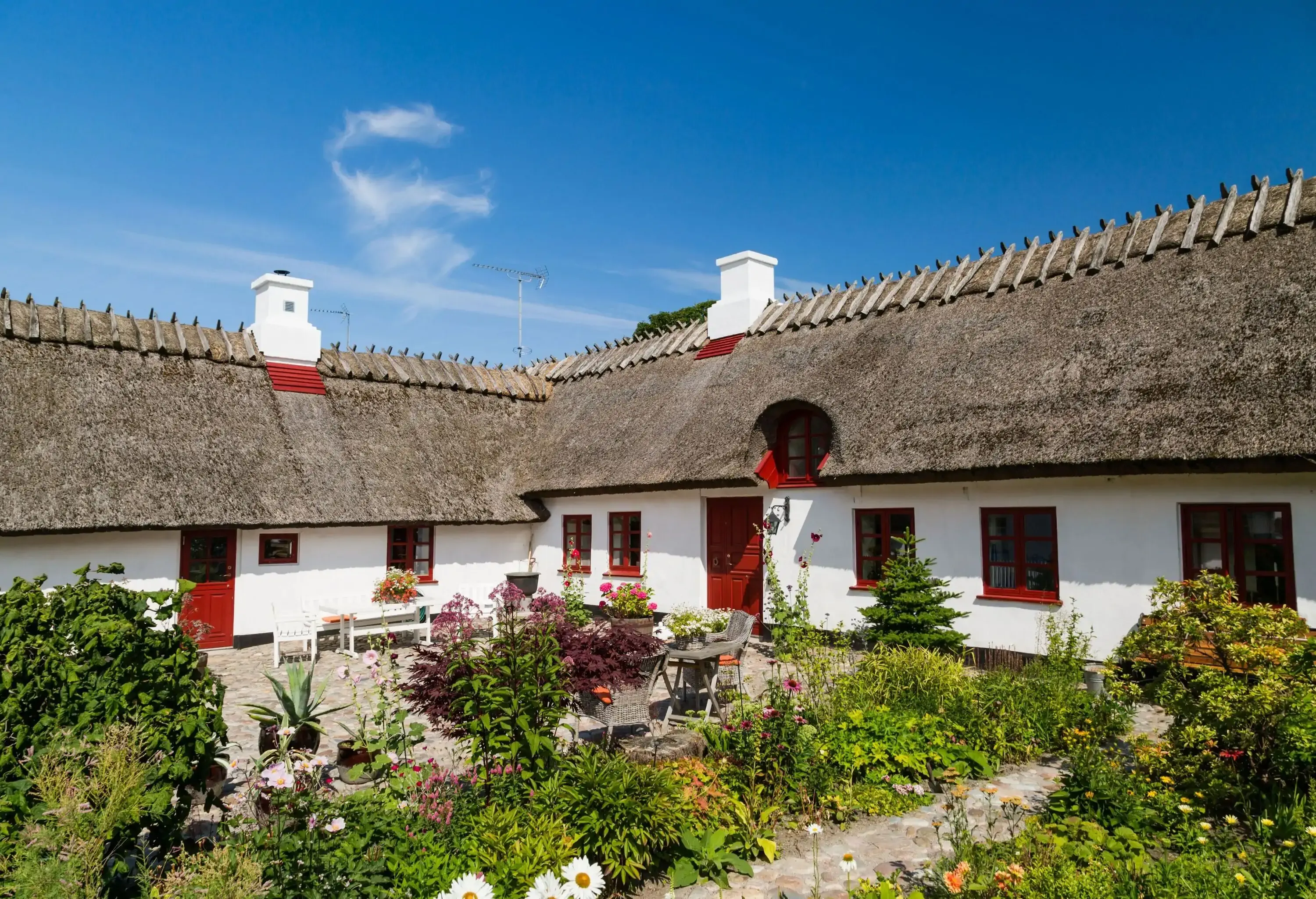 A white farmhouse with thatched roofing surrounding a little patio with plants and chairs.