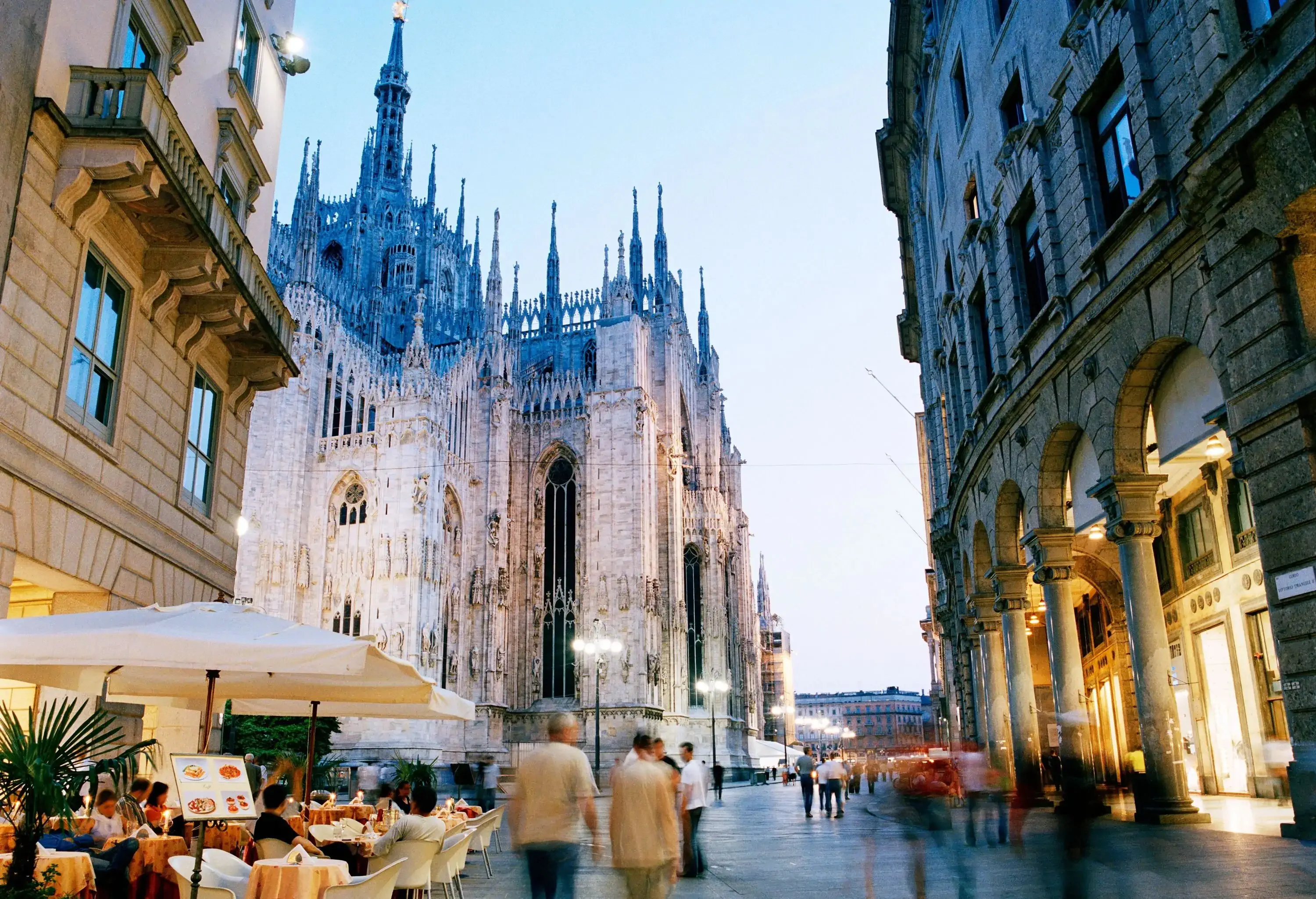 People dine outdoors and walk on a bustling street in the middle of tall buildings.