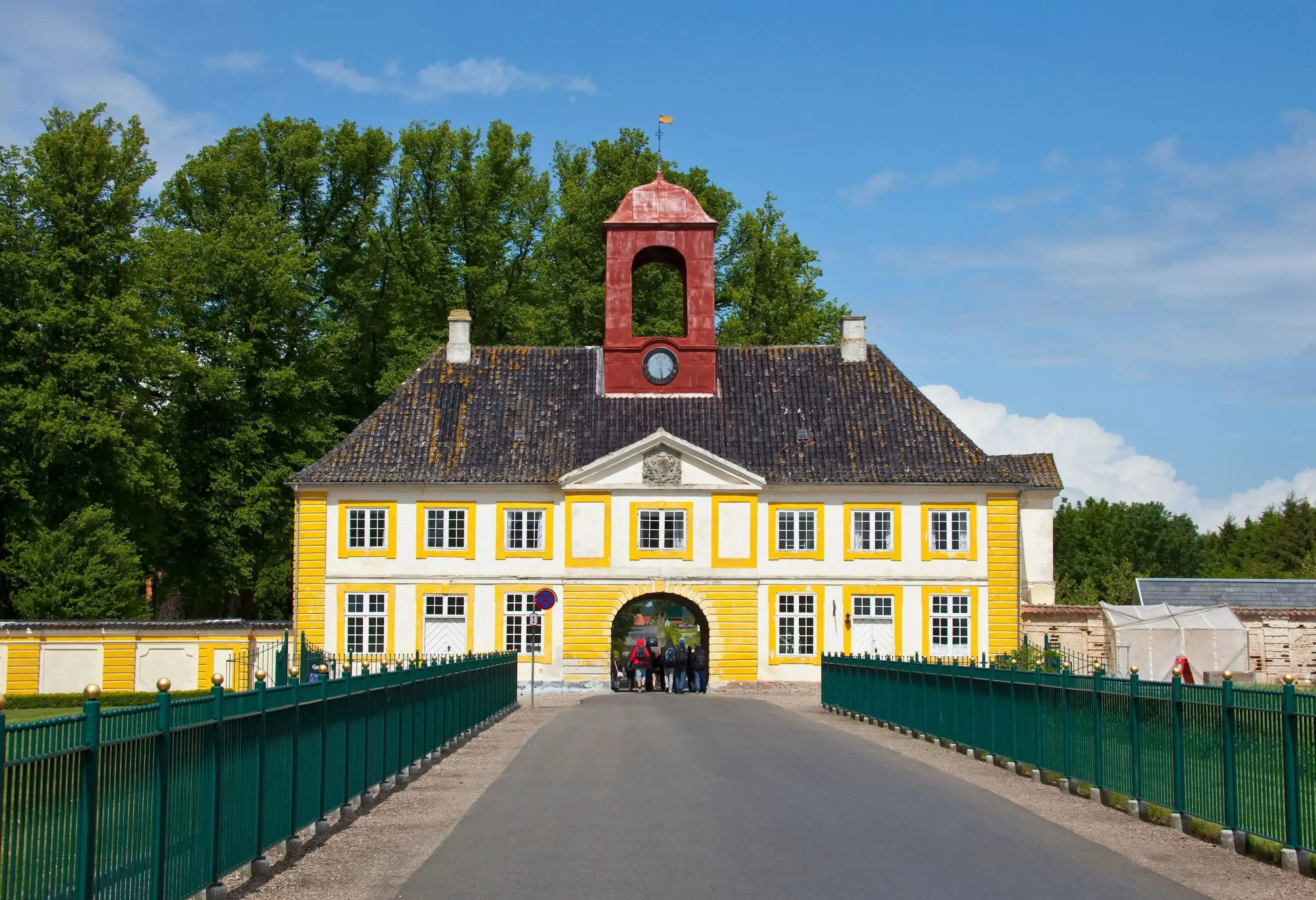 A gated street that leads to a white and yellow building with a central clock tower, an arched entrance, and multiple glass windows.