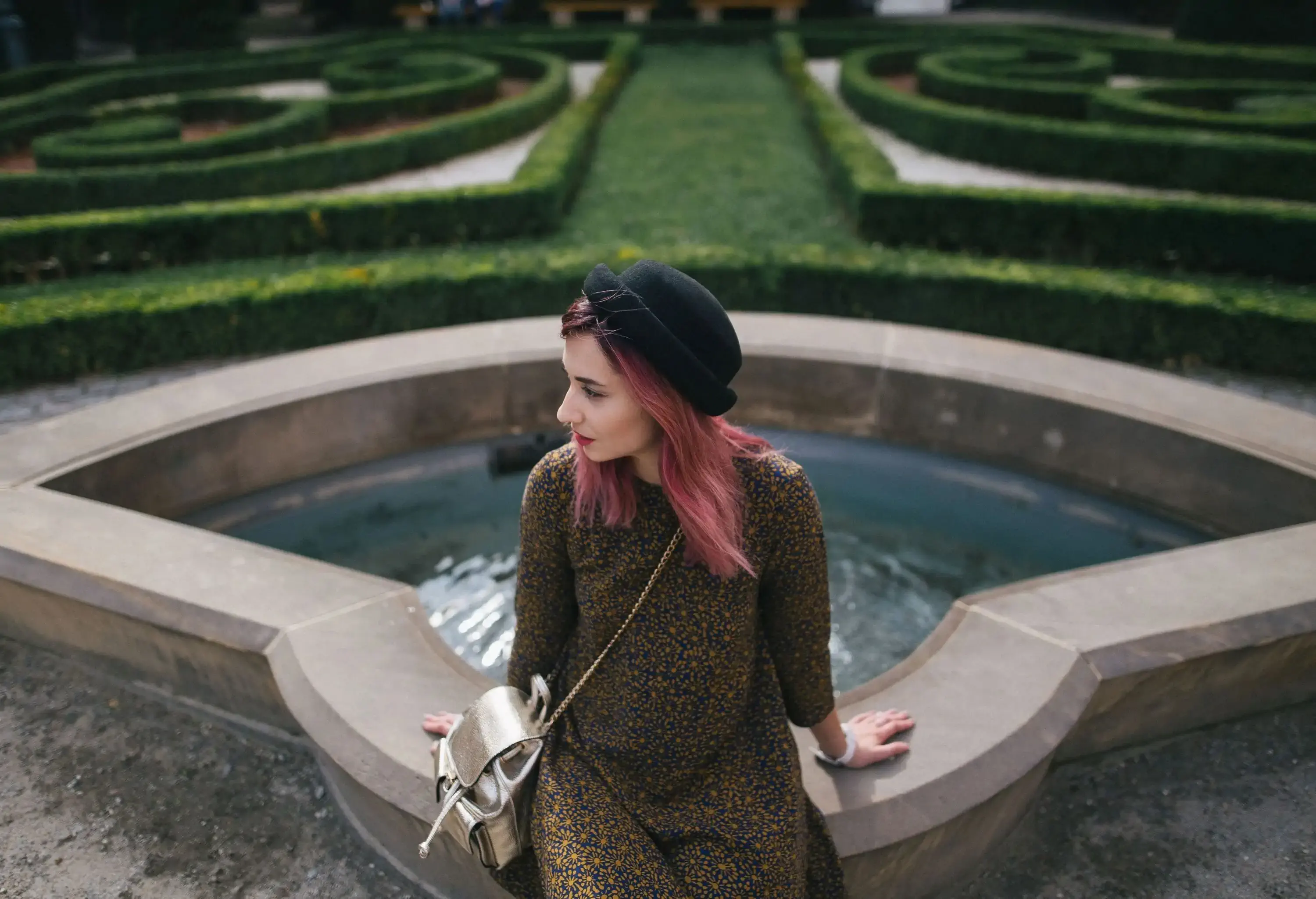 A stylish woman with pinkish hair sits on a fountain in the centre of a maze garden.