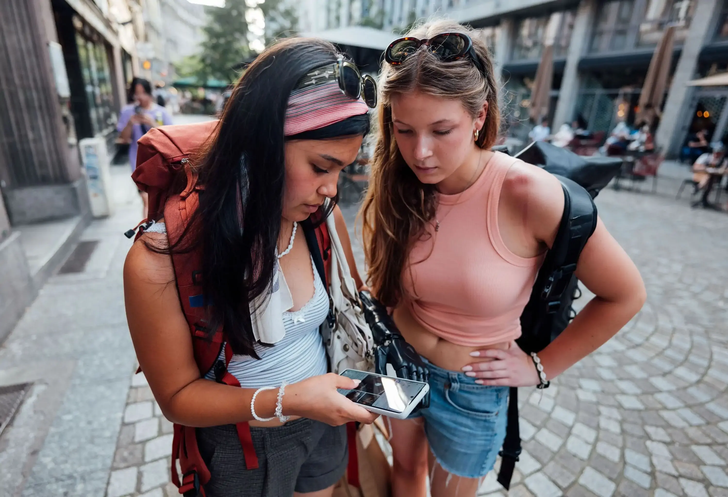 Two women intently looking at a smartphone, with buildings and pedestrians in the background.