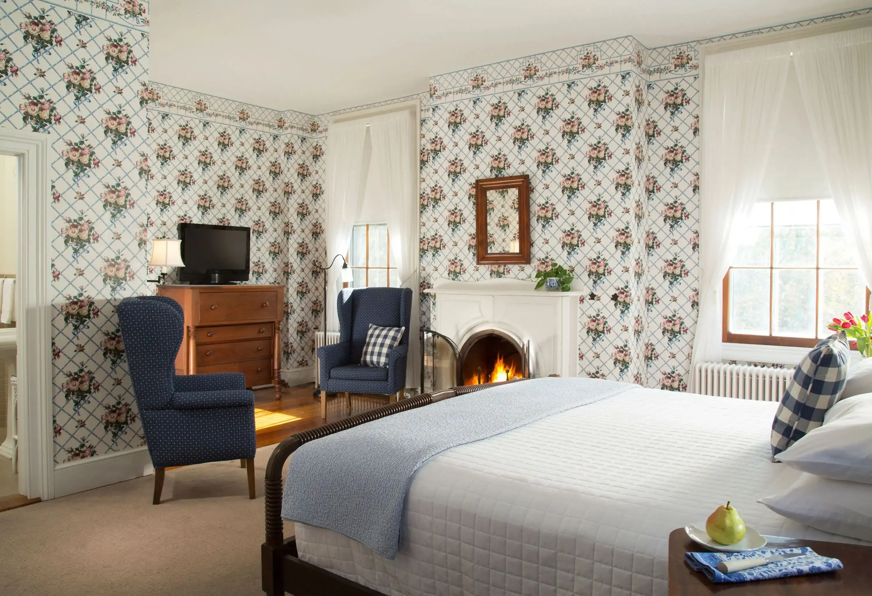 A tidy relaxing bedroom with a fireplace, floral walls, and two blue chairs near the television.