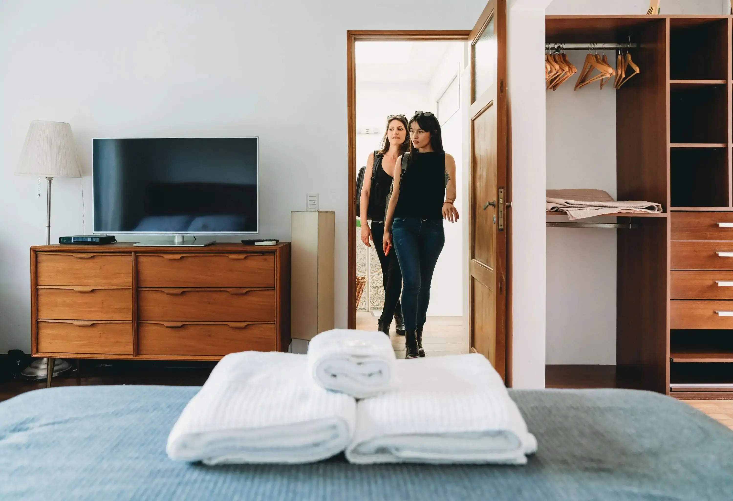 Two women walk into a room with a TV, cabinet, closet, and a bed with fresh towels.