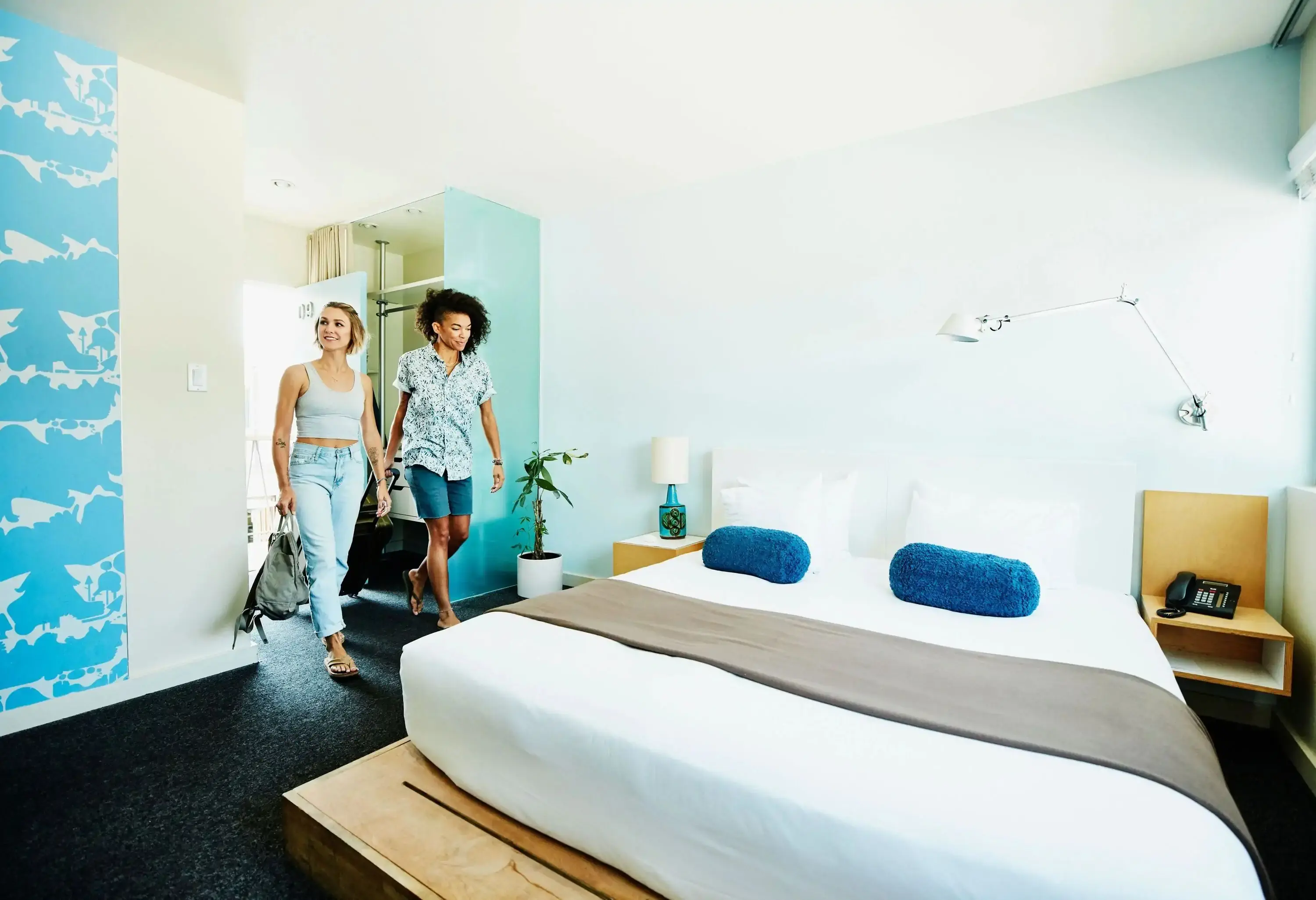 A couple enters a room with a white bed with blue towels and a cabinet.