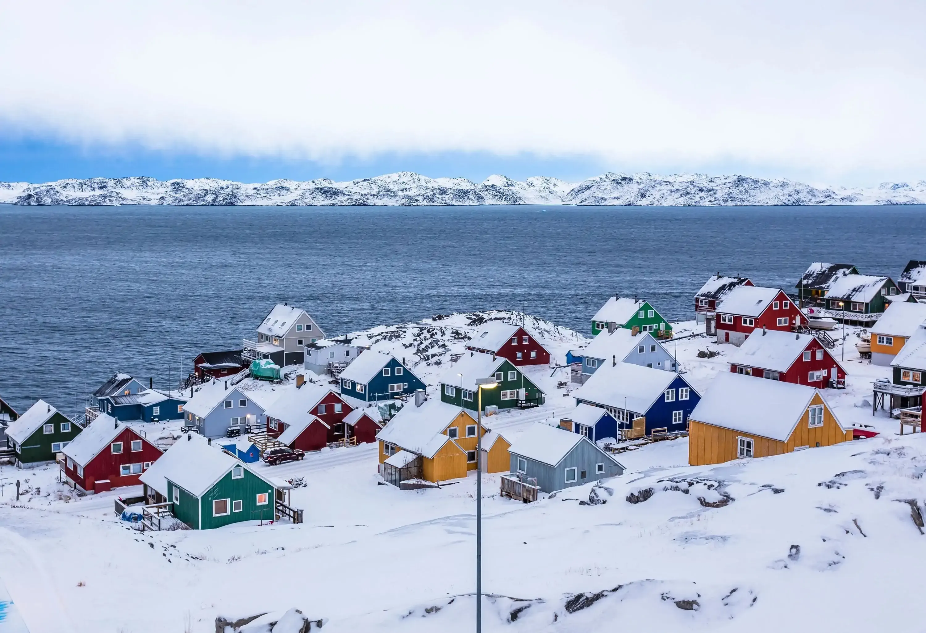 Colourful Inuit houses nestled among rocky terrain and snow-covered surroundings, situated by a fjord in a suburban area.