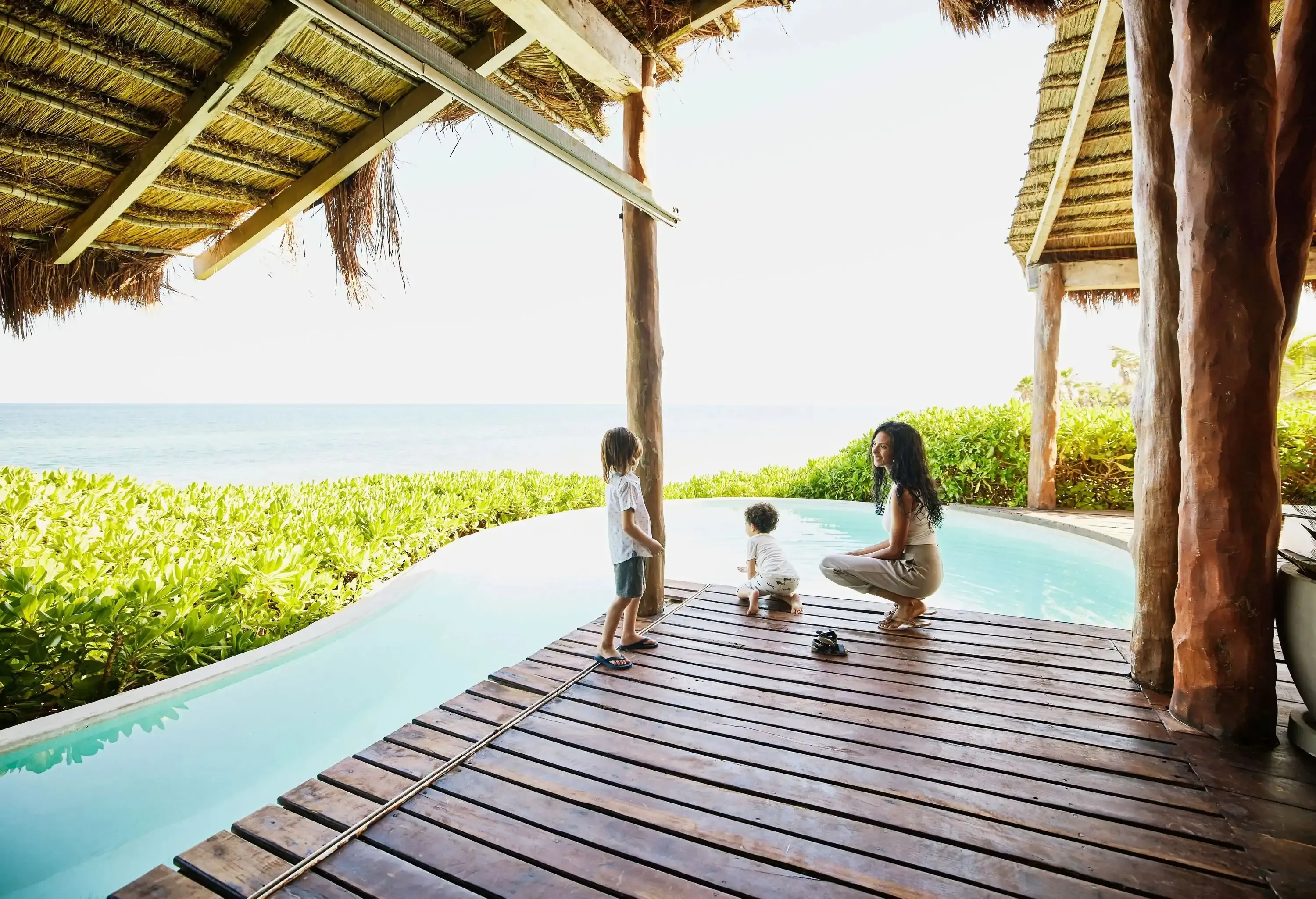 A mother and her two kids play on the wooden platform alongside the swimming pool overlooking the ocean.