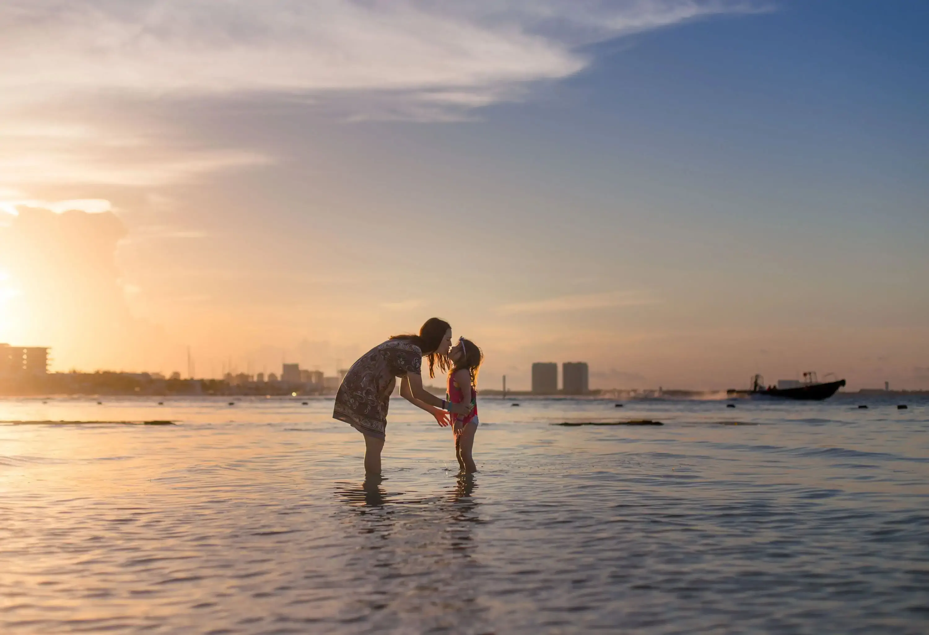 A woman kisses a kid while standing on a beach's shallow waters.