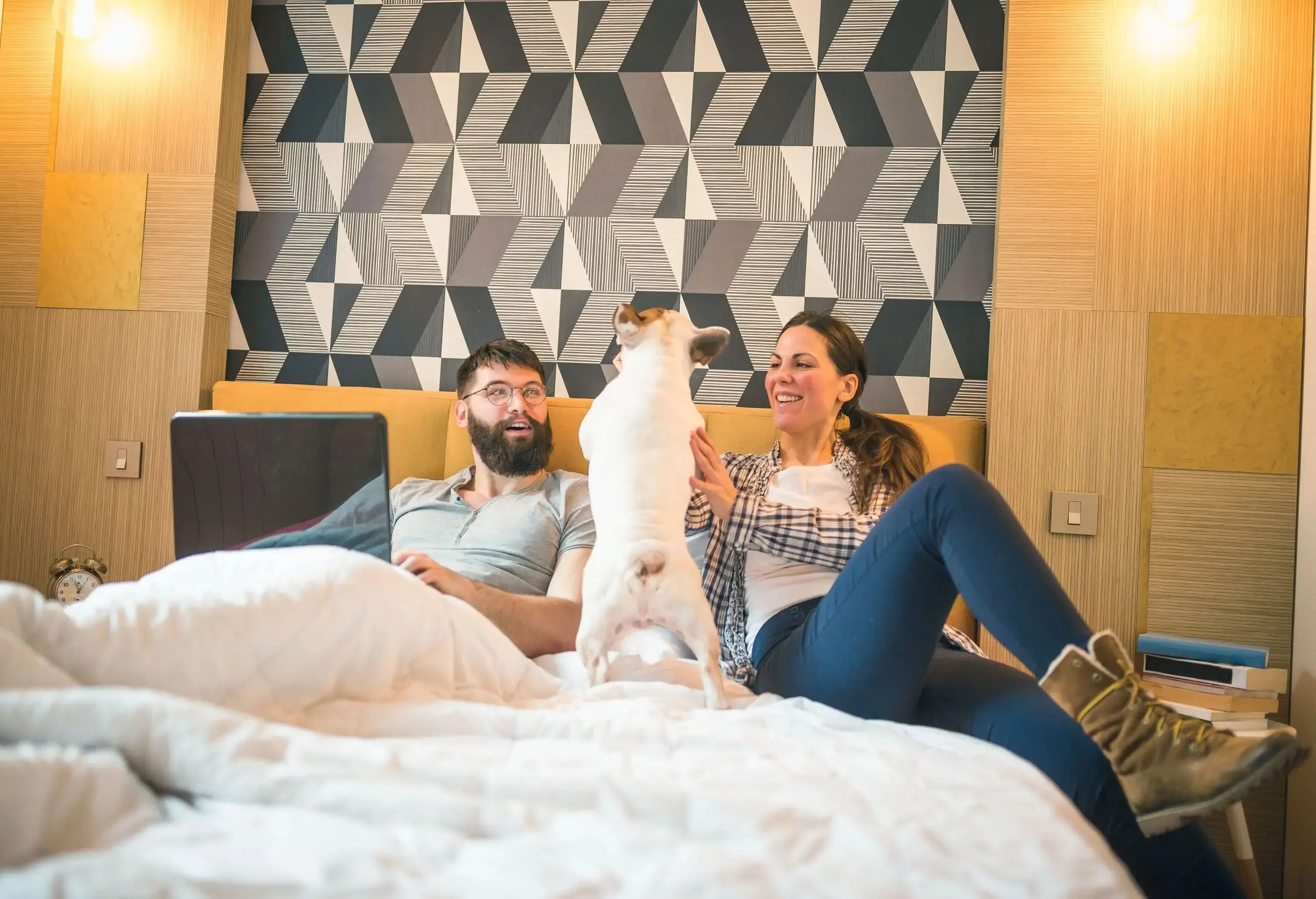 A couple is seated on their bed, with the woman stroking their dog and the man using a laptop.