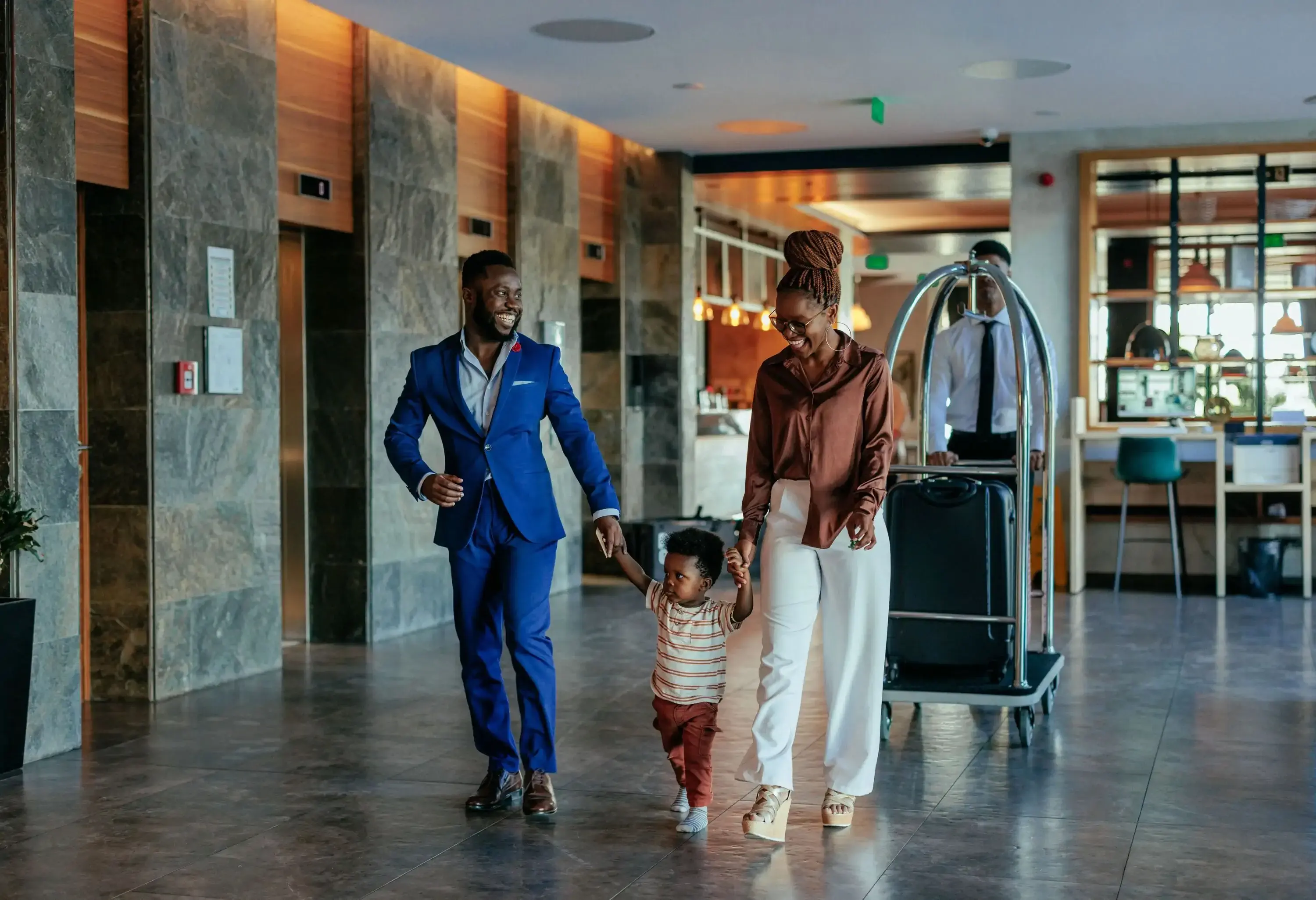 An African American family is walking through the lobby of a luxurious resort. They are fashionably dressed and the hotel attendant is walking behind them pushing the luggage cart.