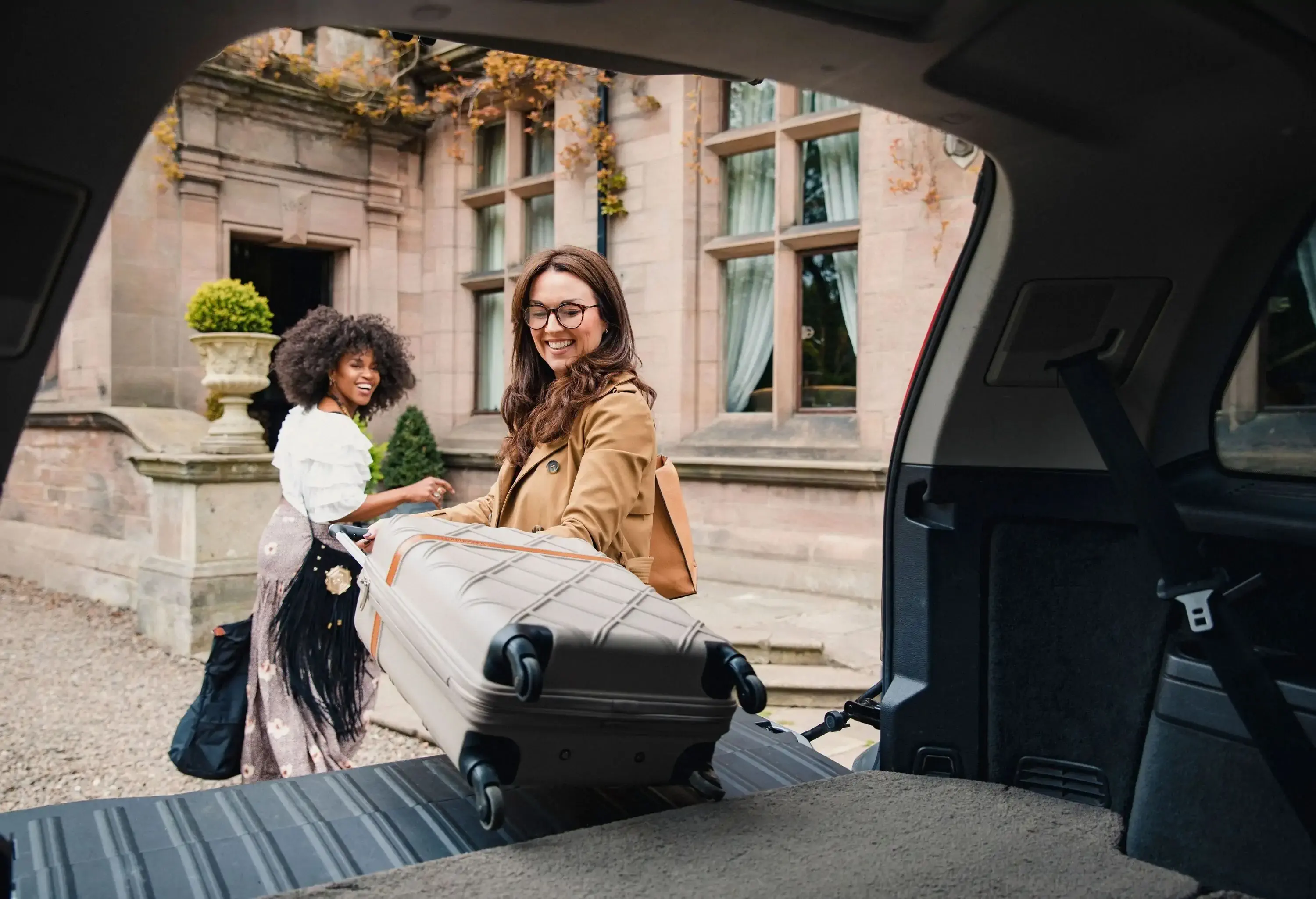 A smiling woman, with another woman beside her, unloading a suitcase from a vehicle in front of an old-fashioned building.