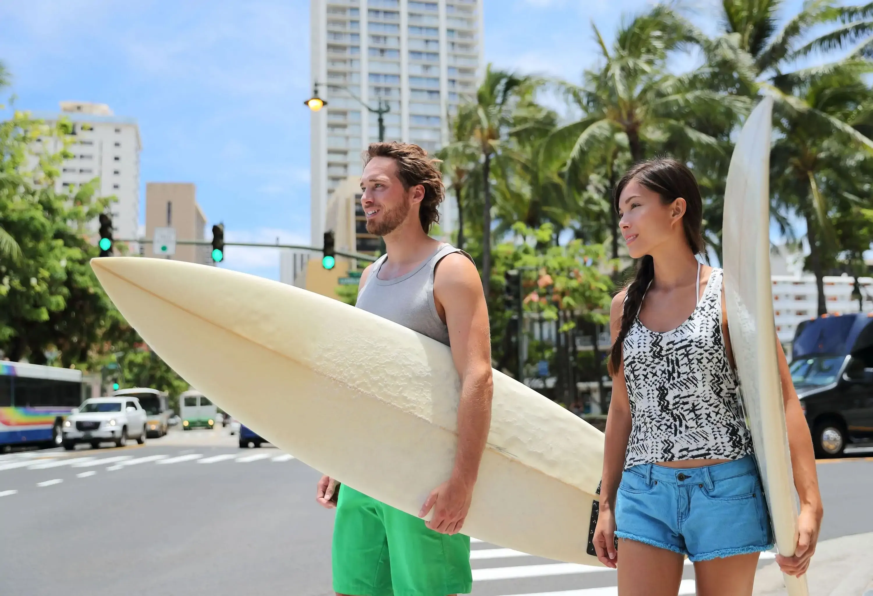 Two people crossing an urban street while carrying surfboards.