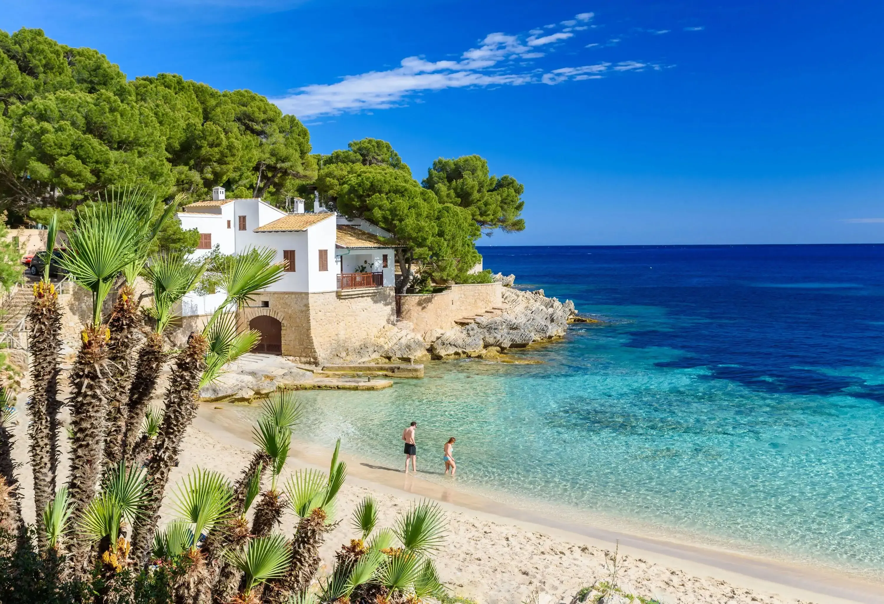 Two people bathe in the shallow waters of a beautiful beach beside a white building located on a rocky coast.