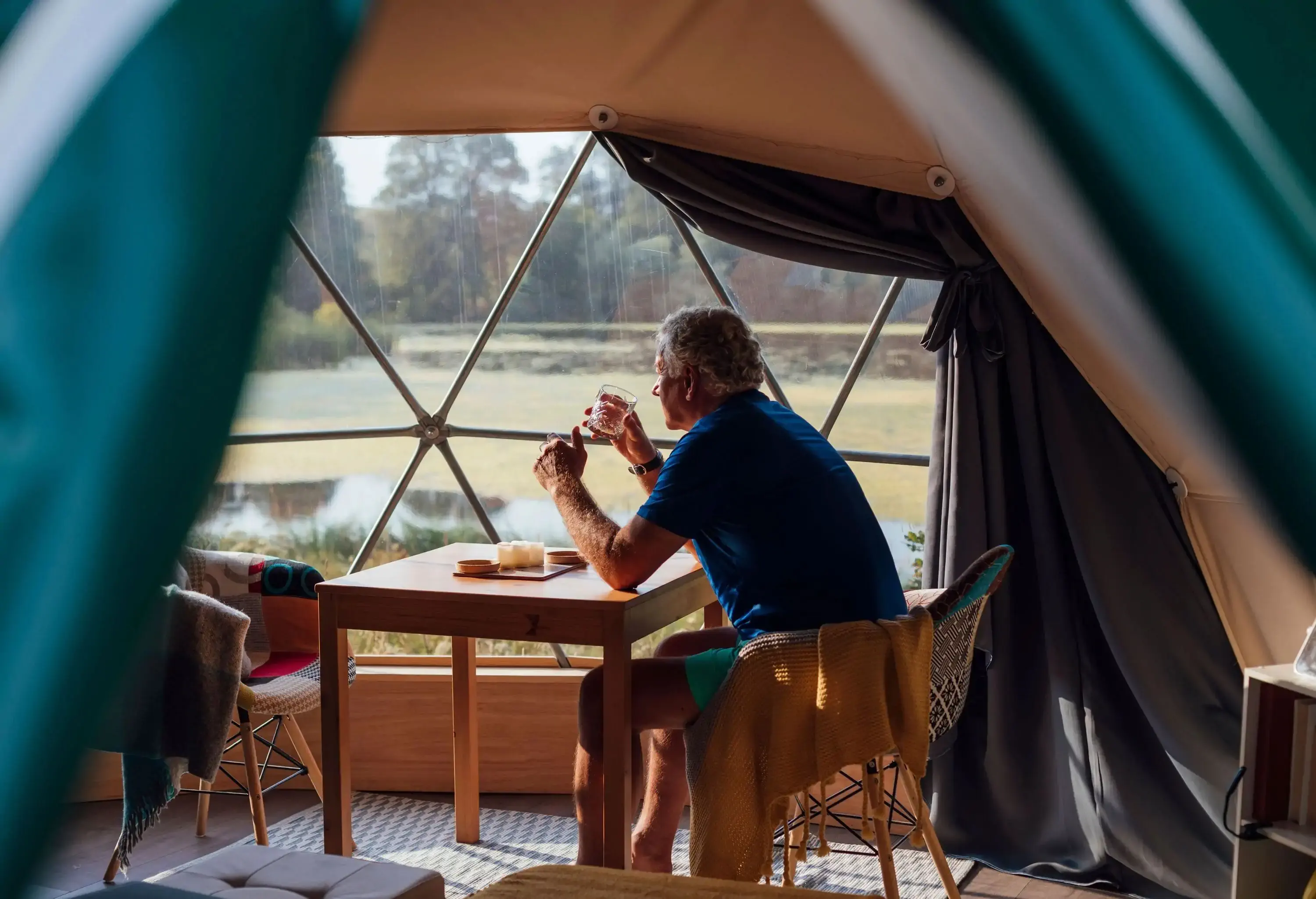 A senior man sitting inside a glamping tent looking through the window with a glass of drink.