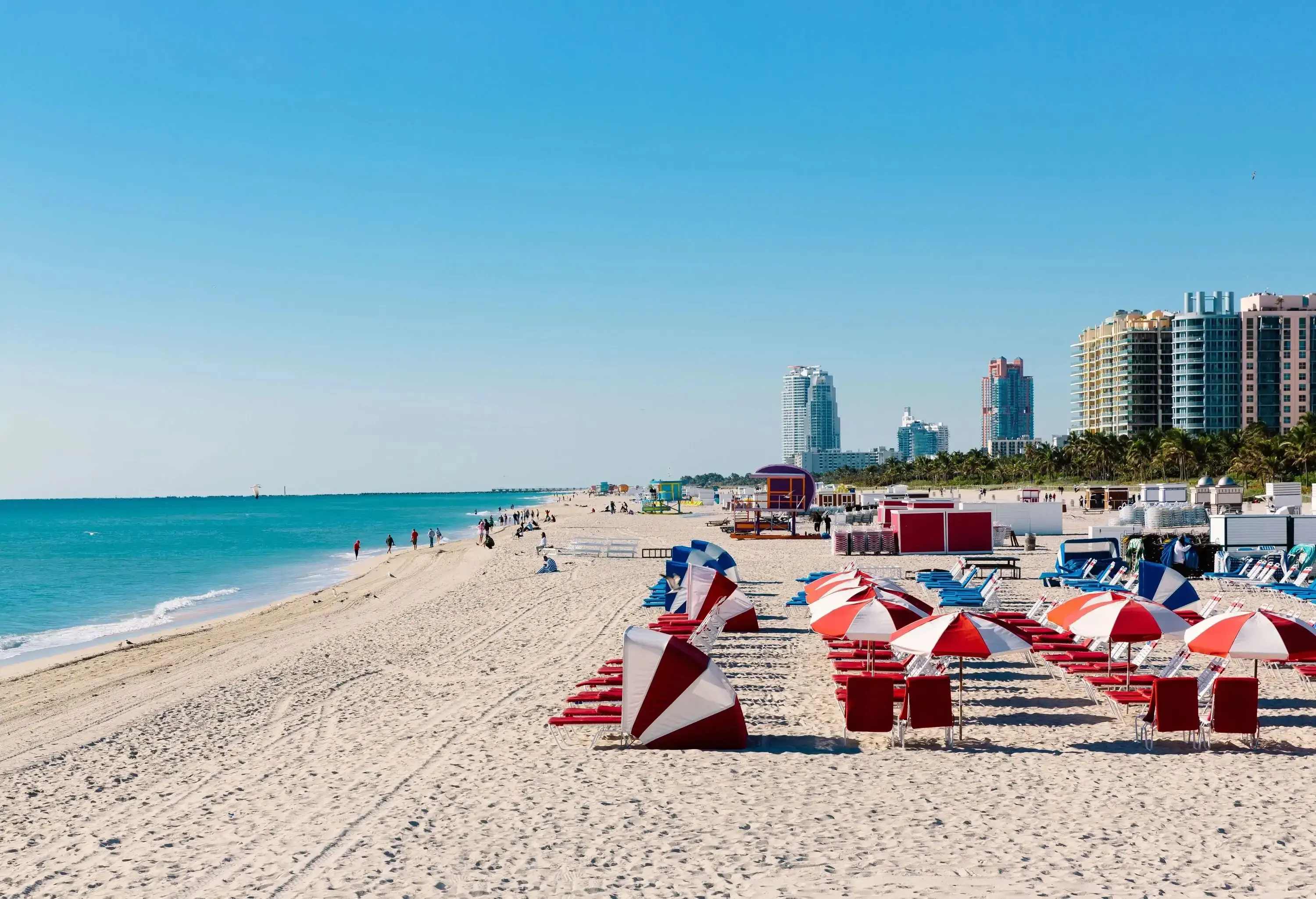 A lively beach features red beach beds nestled under colourful parasols, with beachgoers enjoying the water in the distance and an impressive skyline of towering buildings on the horizon.