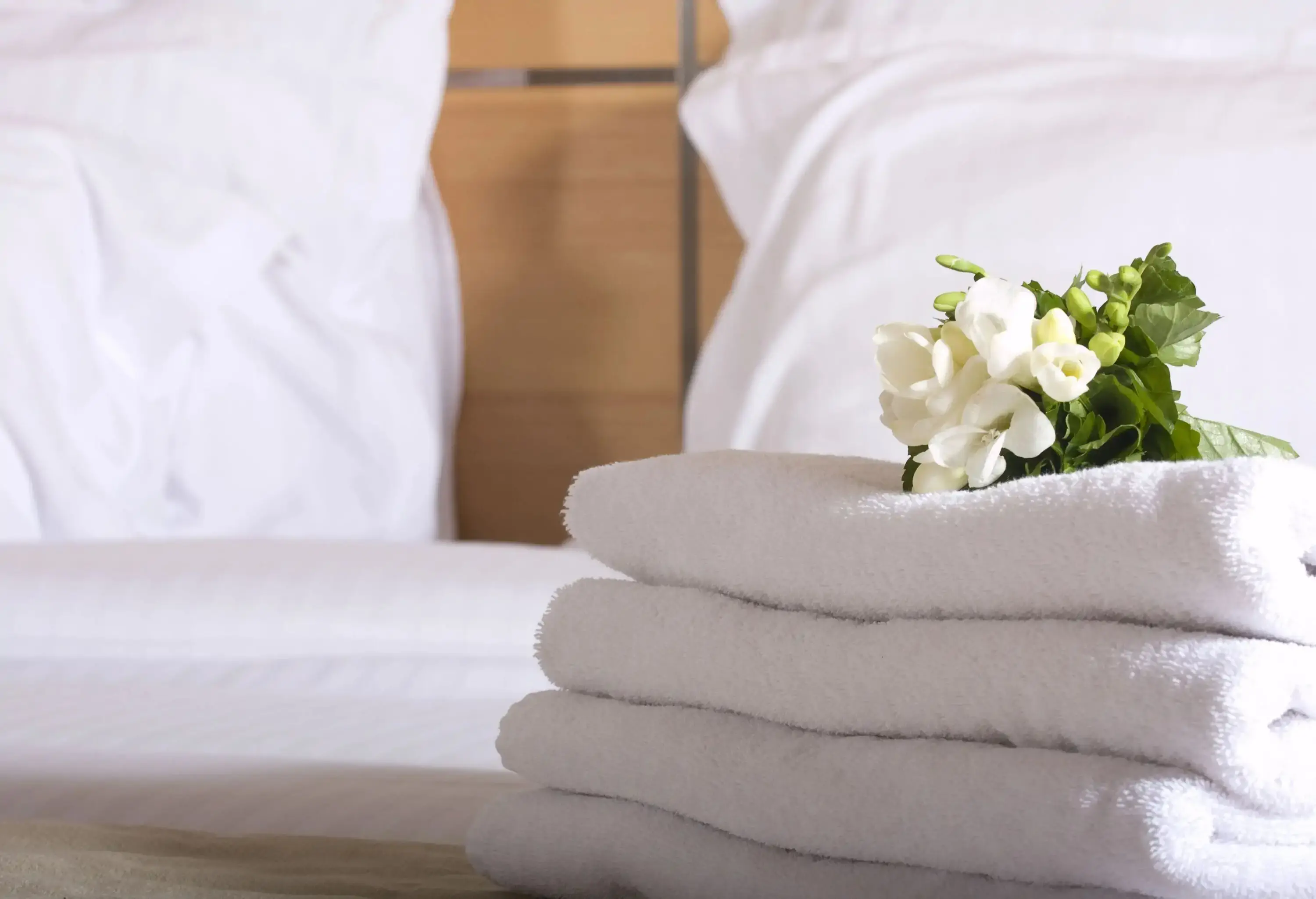 A hotel bed with a bundle of white flowers on top of a stack of fresh towels.