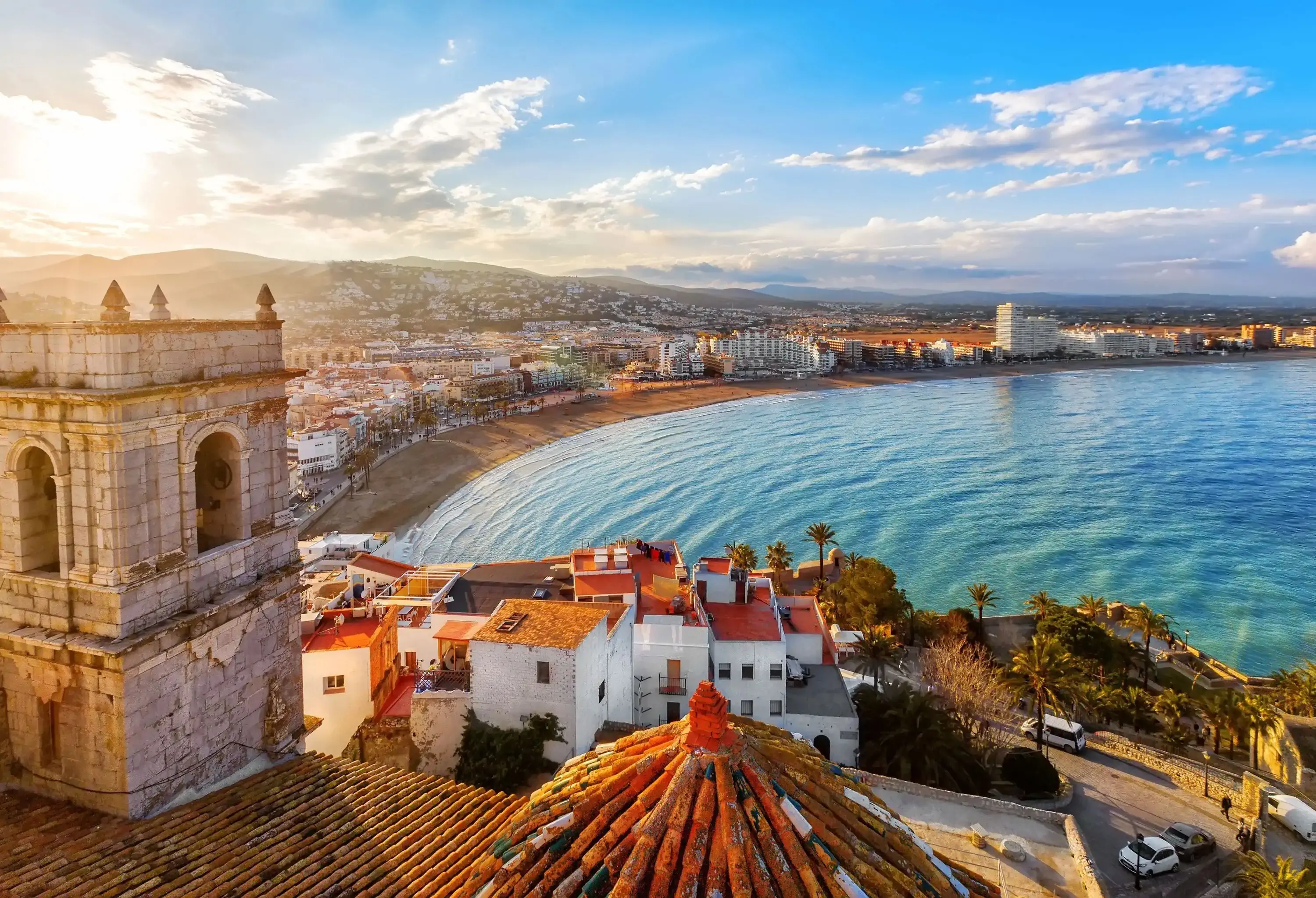 On top of a castle's roof is a view of a tightly packed city along the sea.