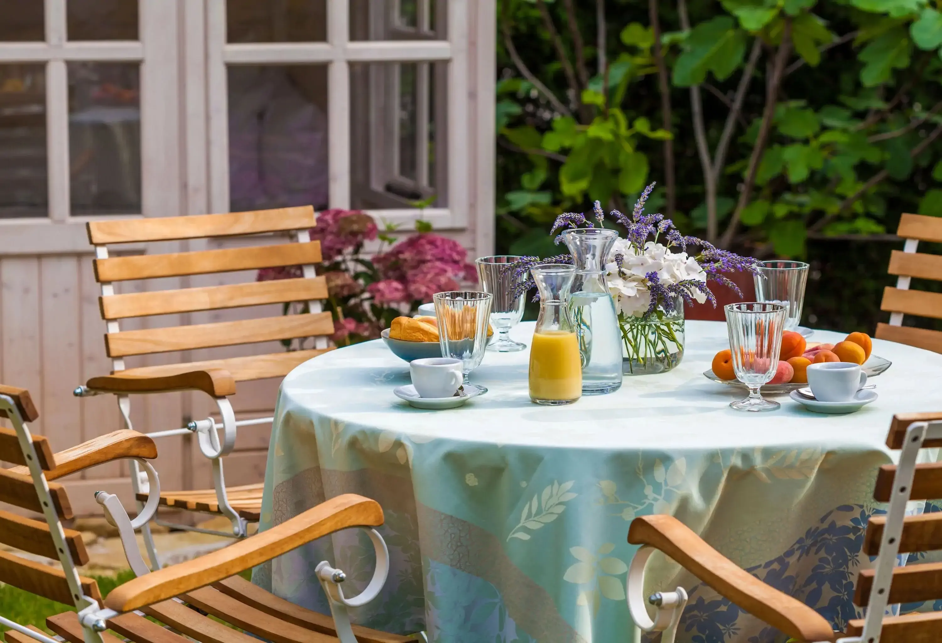 A pitcher of juice, water, fruit, and bread set on a patio table with wooden chairs.