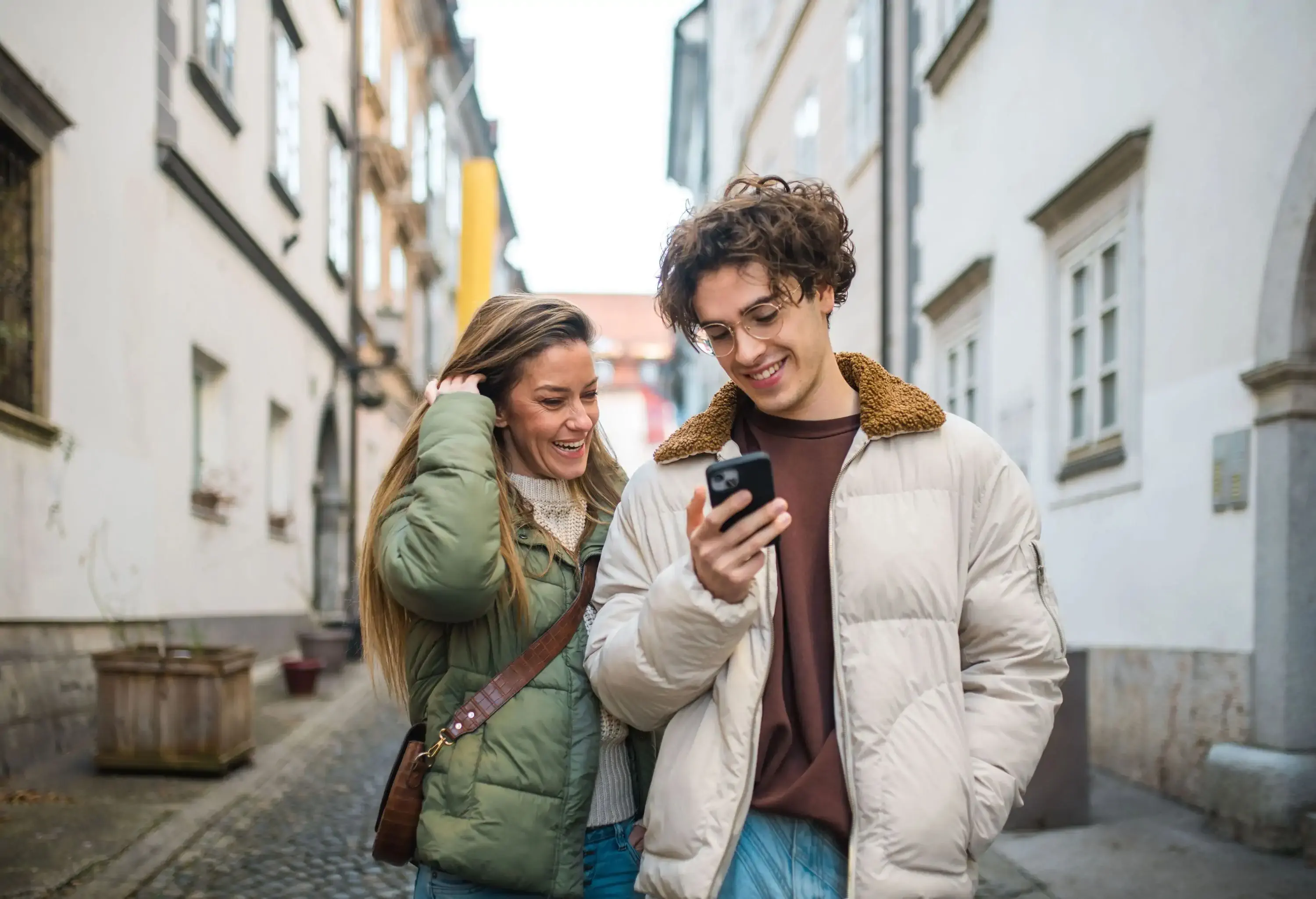 Two smiling people looking at a phone together while standing on a narrow street bordered by buildings.