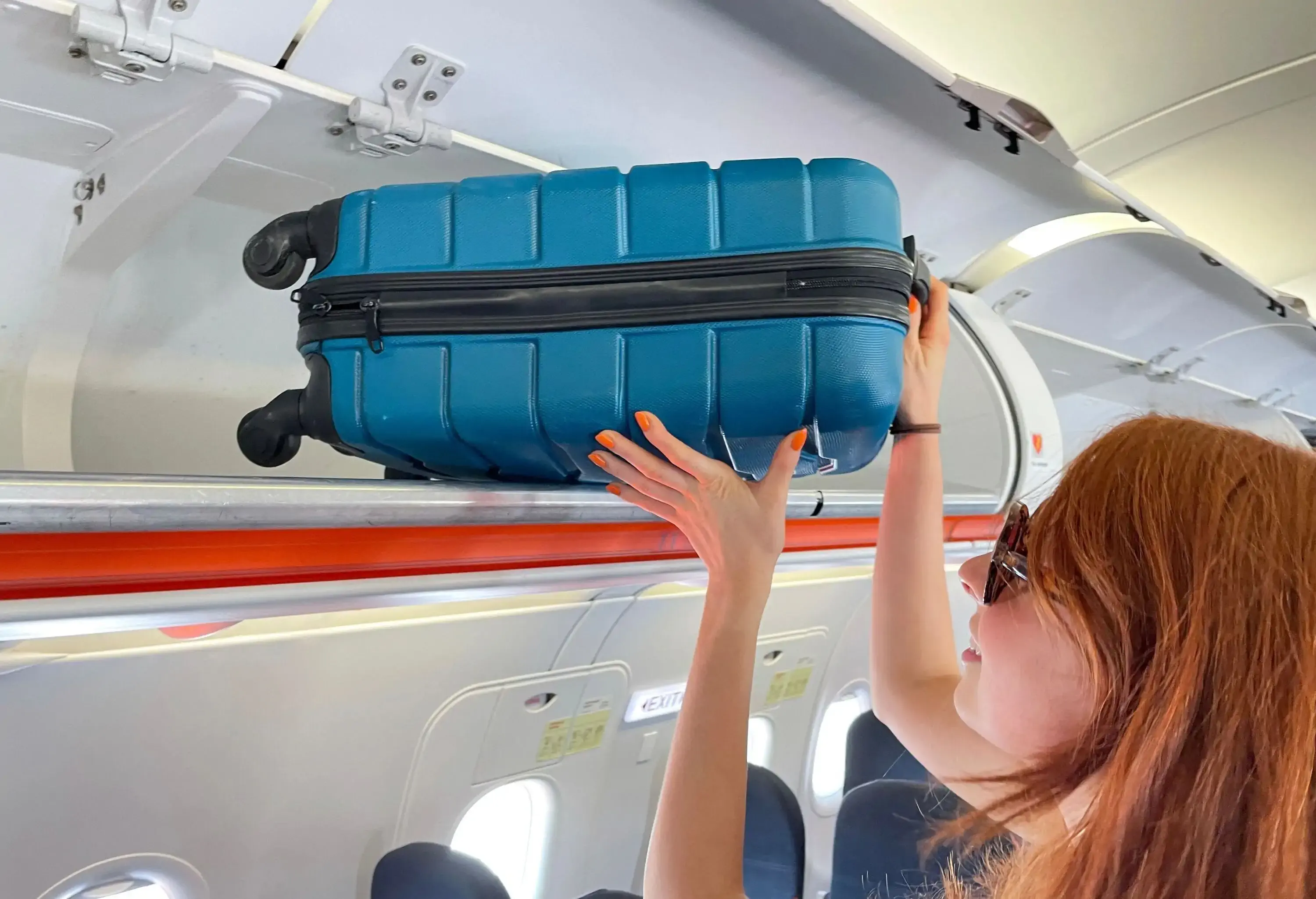 A row of carryon luggage overhead baggage compartments in an airplane cabin, a red haired woman putting away her suitcase.