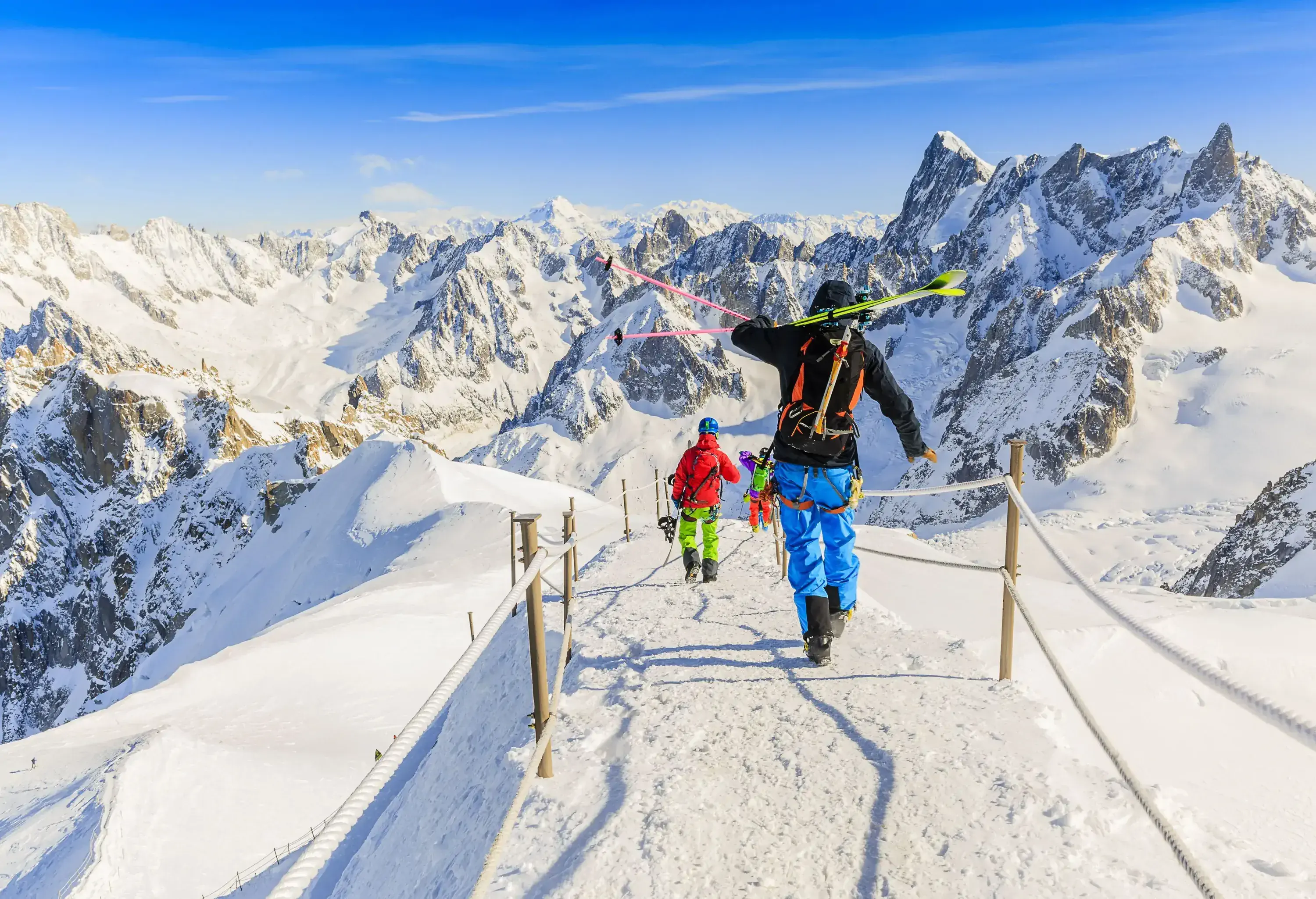 Skiers carrying equipment walking along the ridge of a snow-covered mountain with rope barrier.