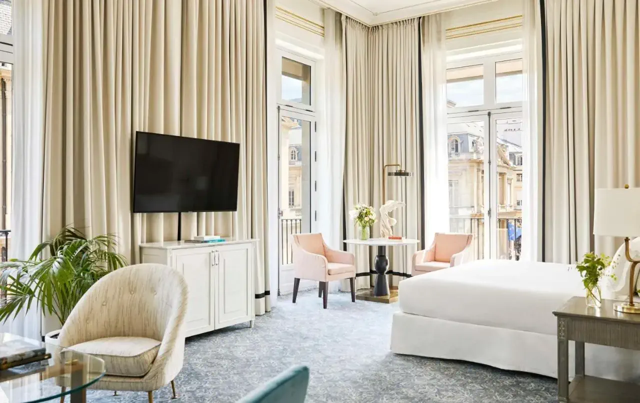 Elegant hotel room with classic decor, featuring a large bed with draped canopy, vintage chandelier, ornate curtains, and a balcony with outdoor seating.