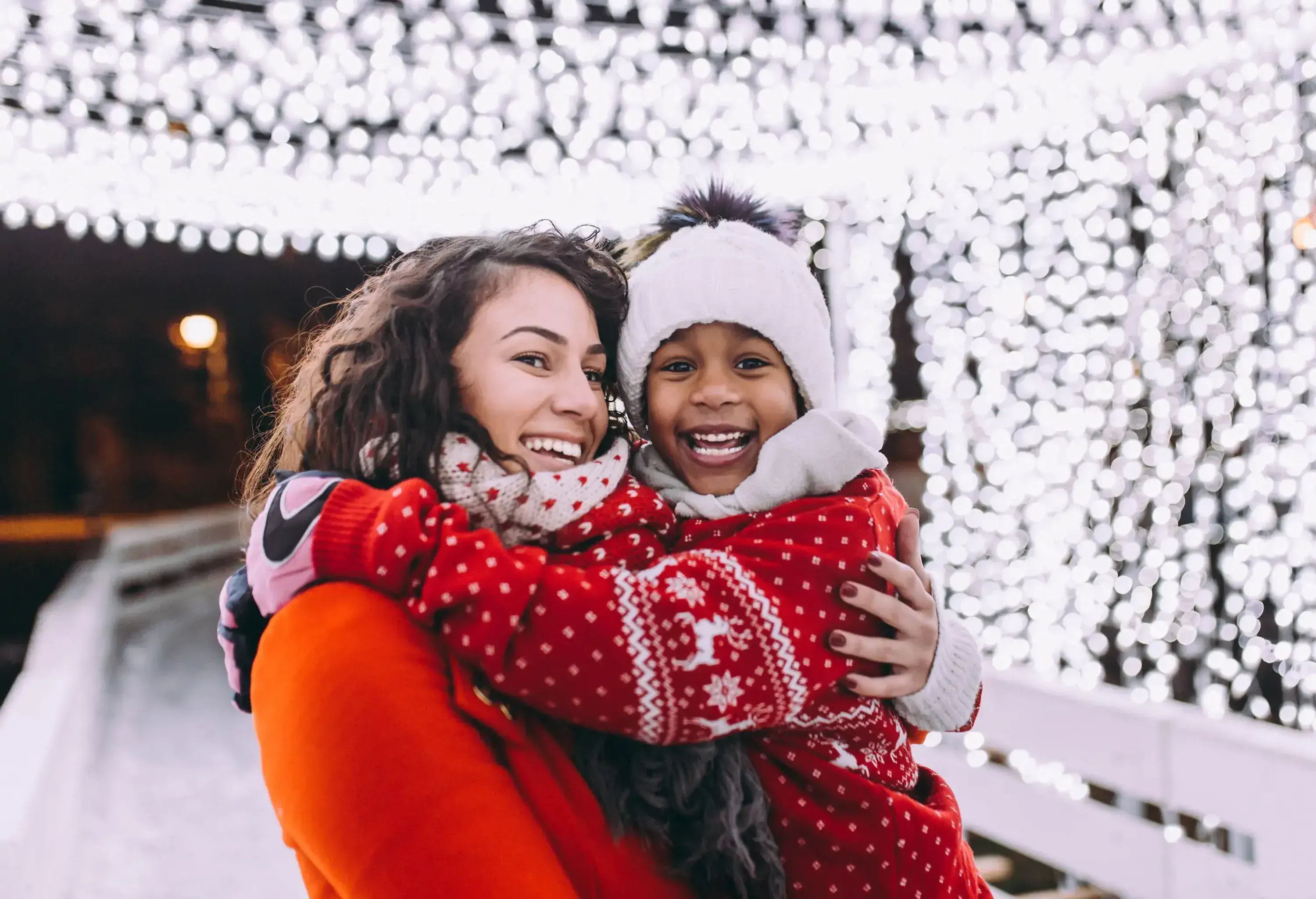 A woman and a young child smiling and holding on to each other as they stand under a canopy of lights.