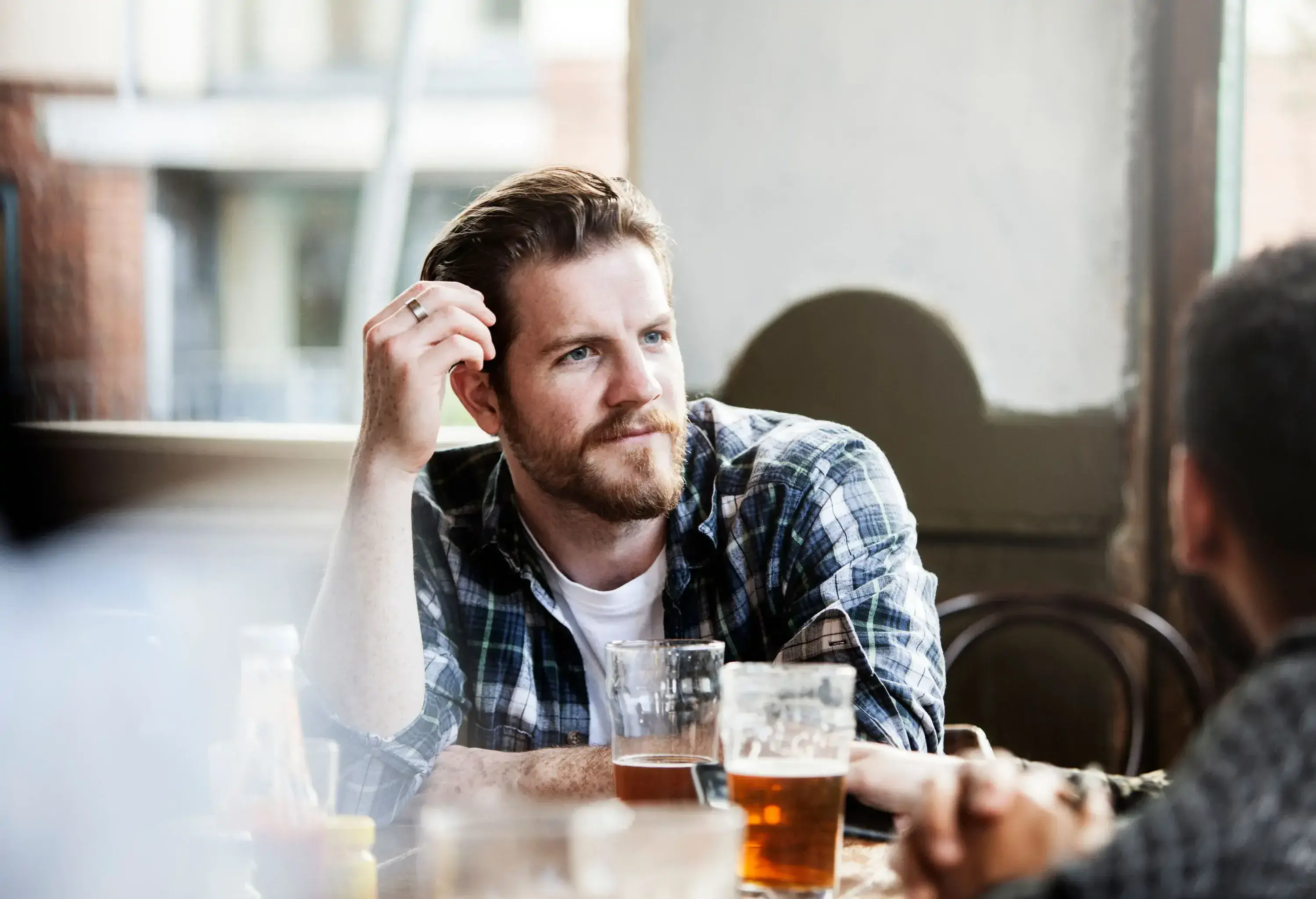 A bearded man in a chequered shirt listens to a friend inside a pub.
