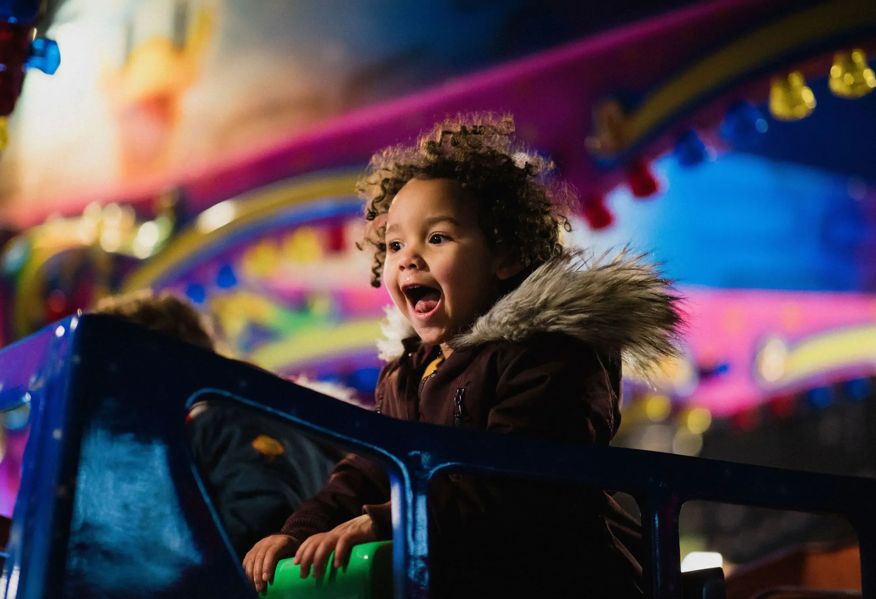 A curly-haired kid in a fur coat enjoys riding a carousel.