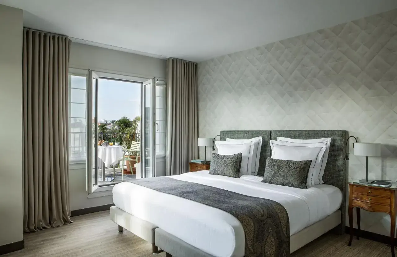 Elegant hotel room with a king-sized bed, plush pillows, and a textured accent wall. Open doors lead to a balcony with a table and chairs, creating a serene atmosphere.