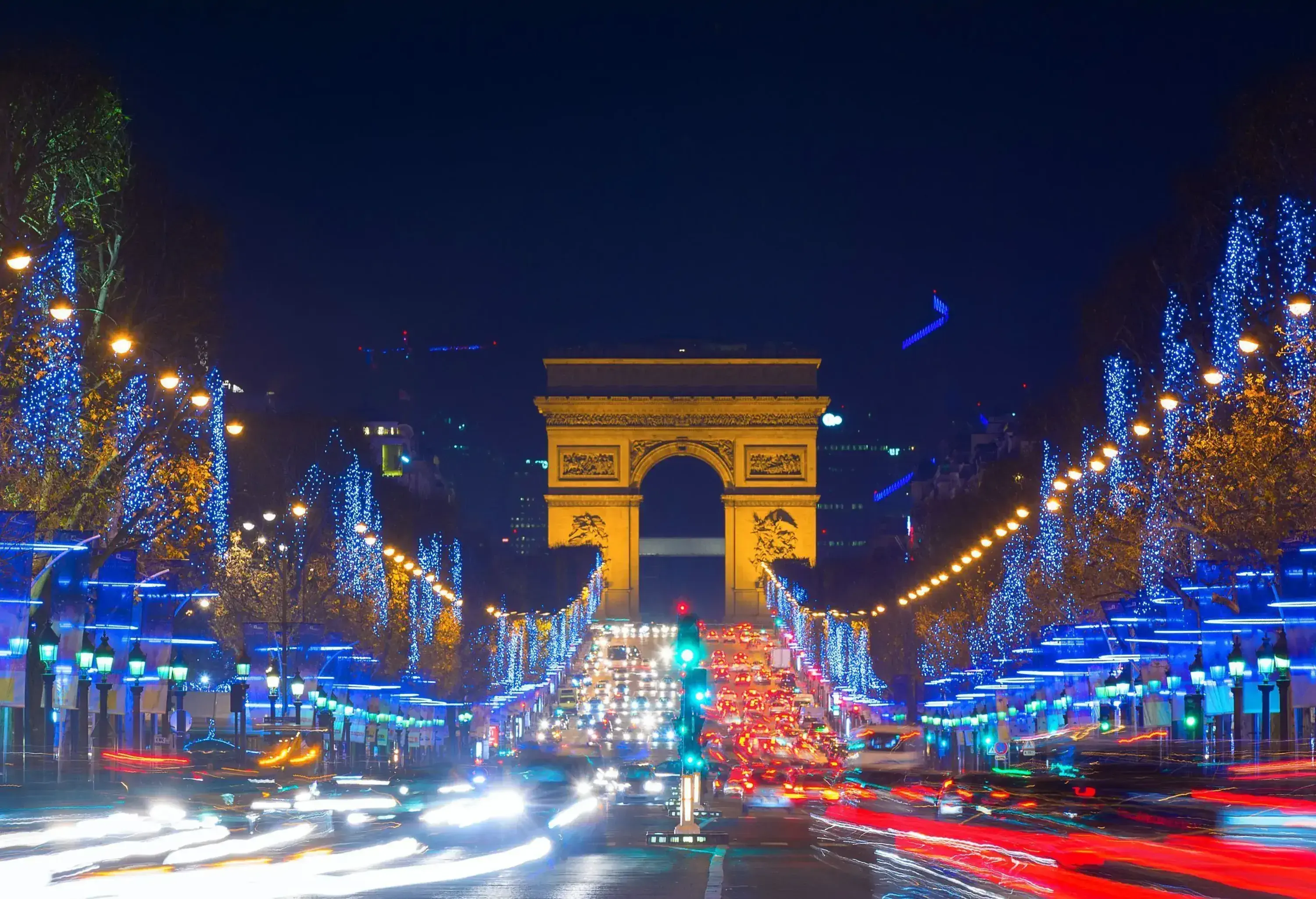 A bustling broad avenue bordered by trees decorated with Christmas lights on both sides heading towards a tall triumphal arch.