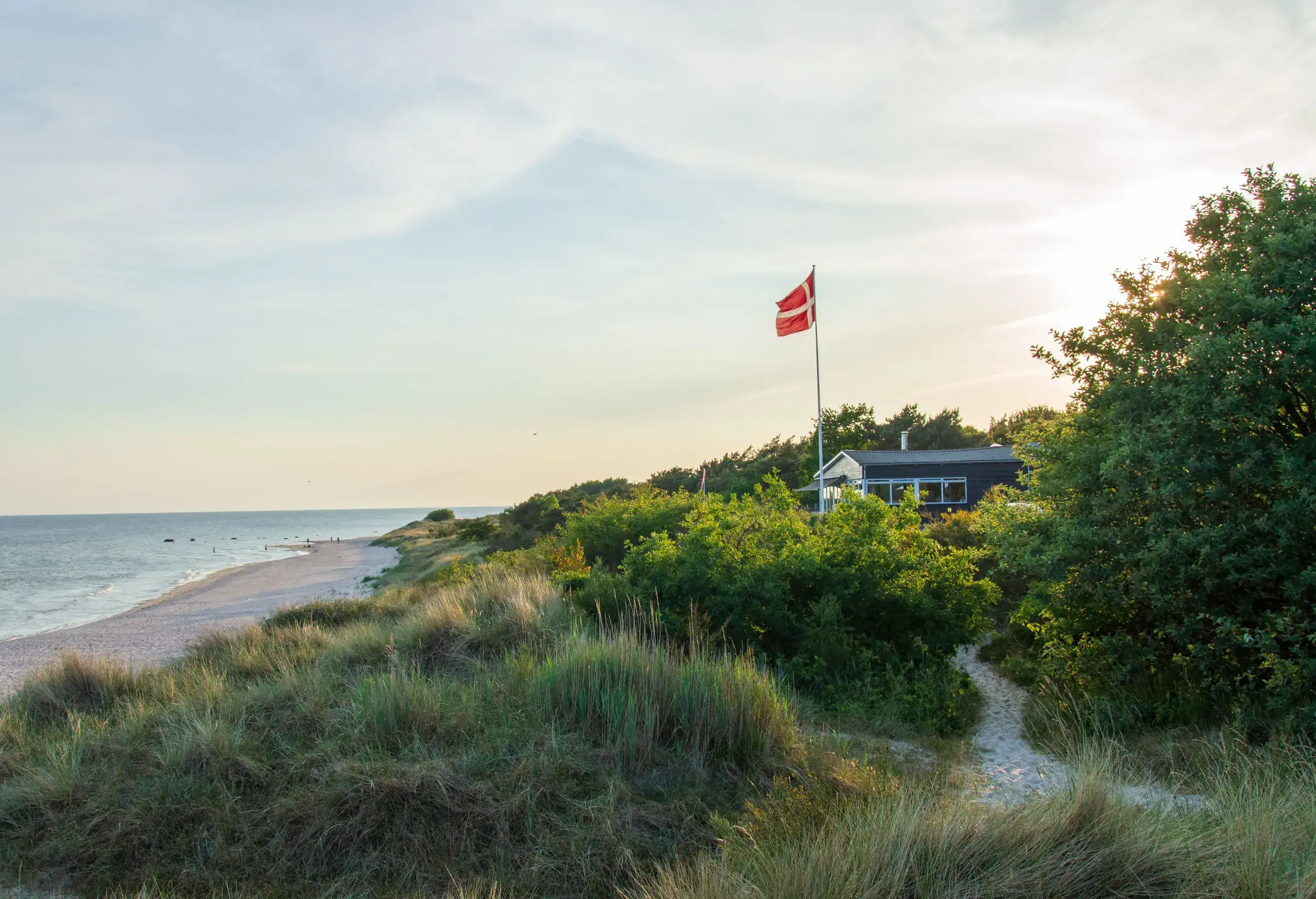 A coastal landscape with a sandy beach and grassy dunes leading to a house with a waving Danish flag under a partly cloudy sky.