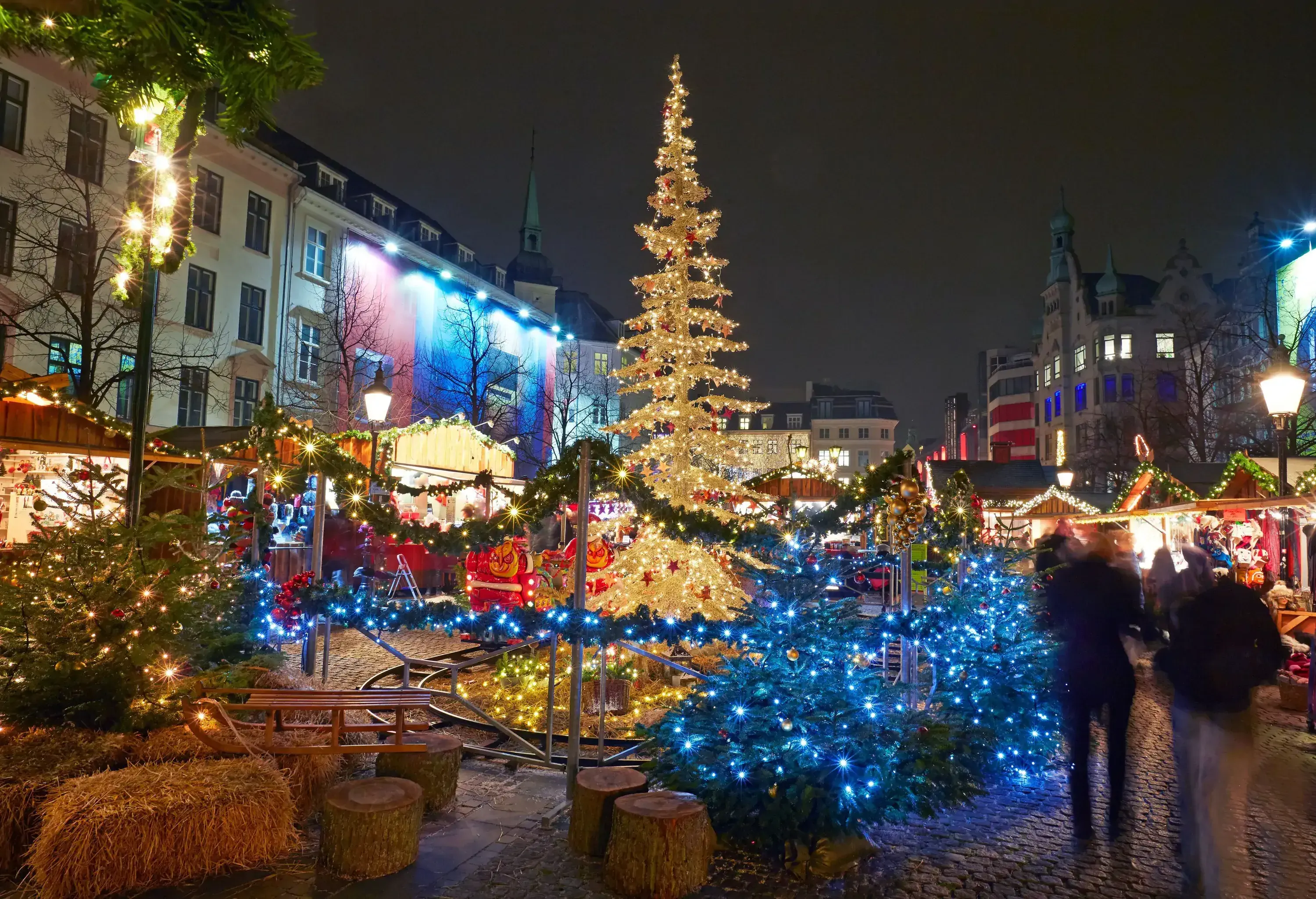 Christmas market at night glowing with warm lights and vibrant decorations amidst city architecture.
