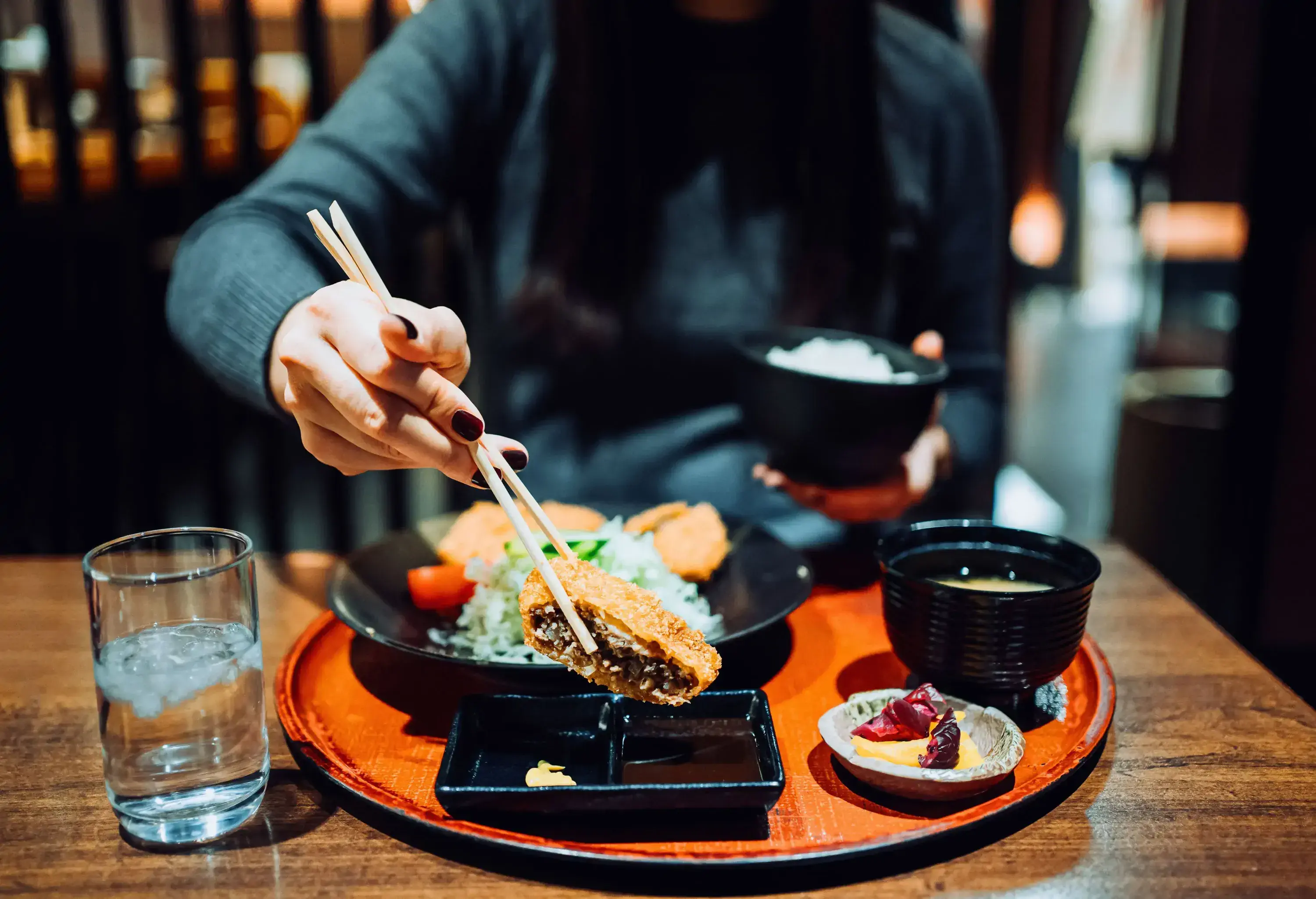 A woman savours a traditional Japanese meal set, complete with miso soup and appetisers, while skillfully holding a piece of crispy tonkatsu with chopsticks, ready to dip into a delectable sauce.