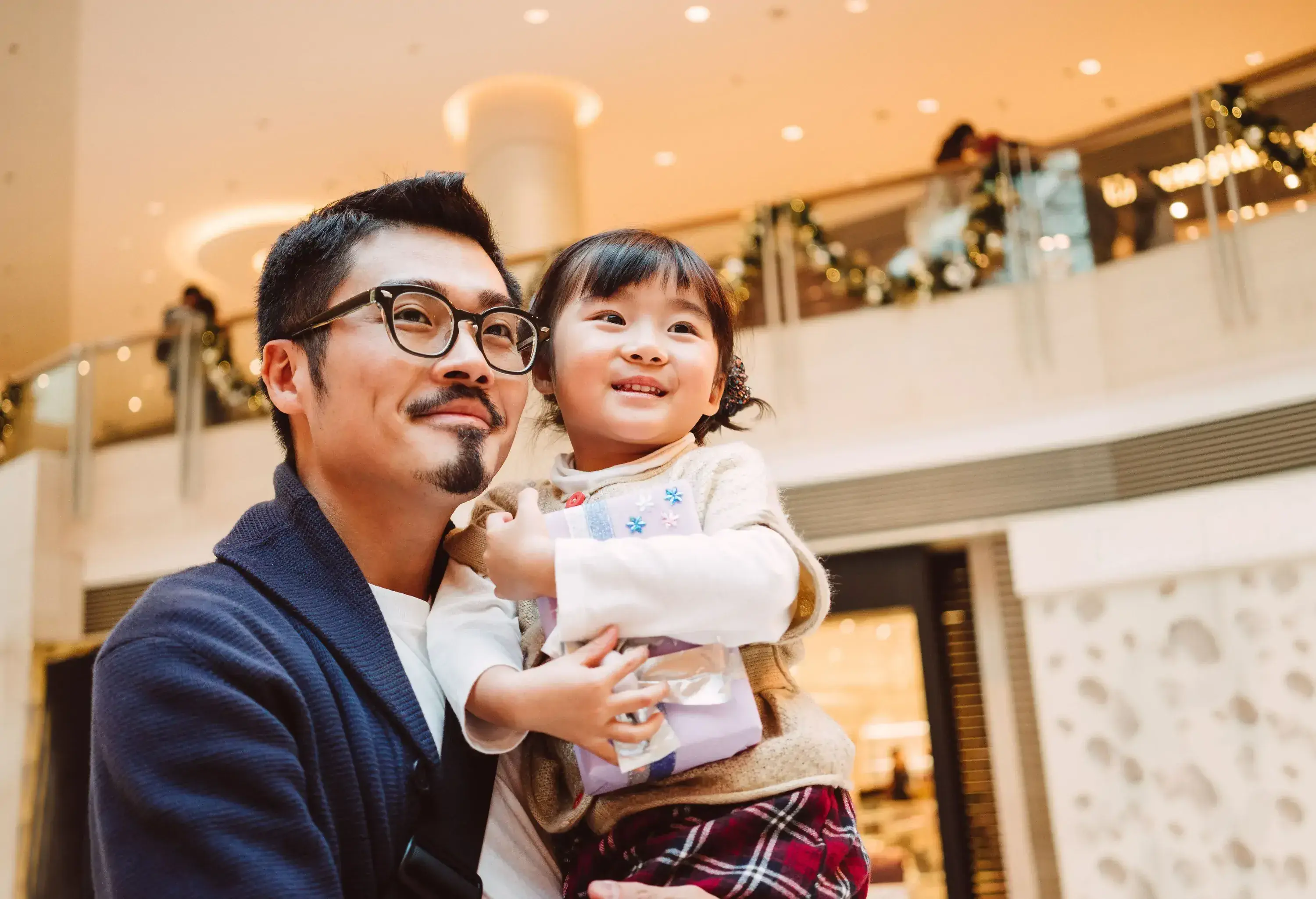 A joyful father carrying his young daughter clutching a wrapped present inside a shopping mall.