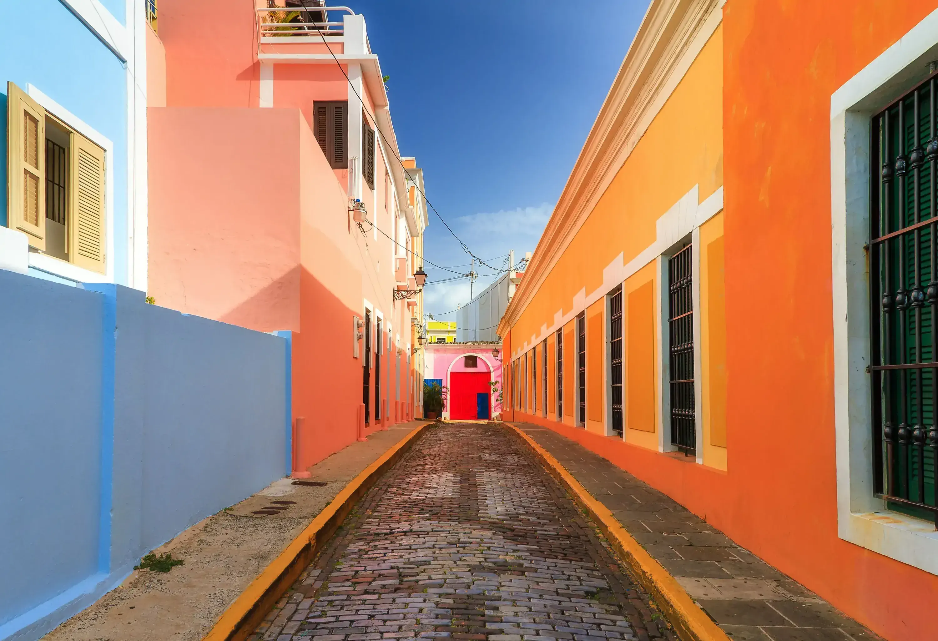 Buildings of various colours lining both sides of a paved alleyway, their rooftops framing the blue sky.