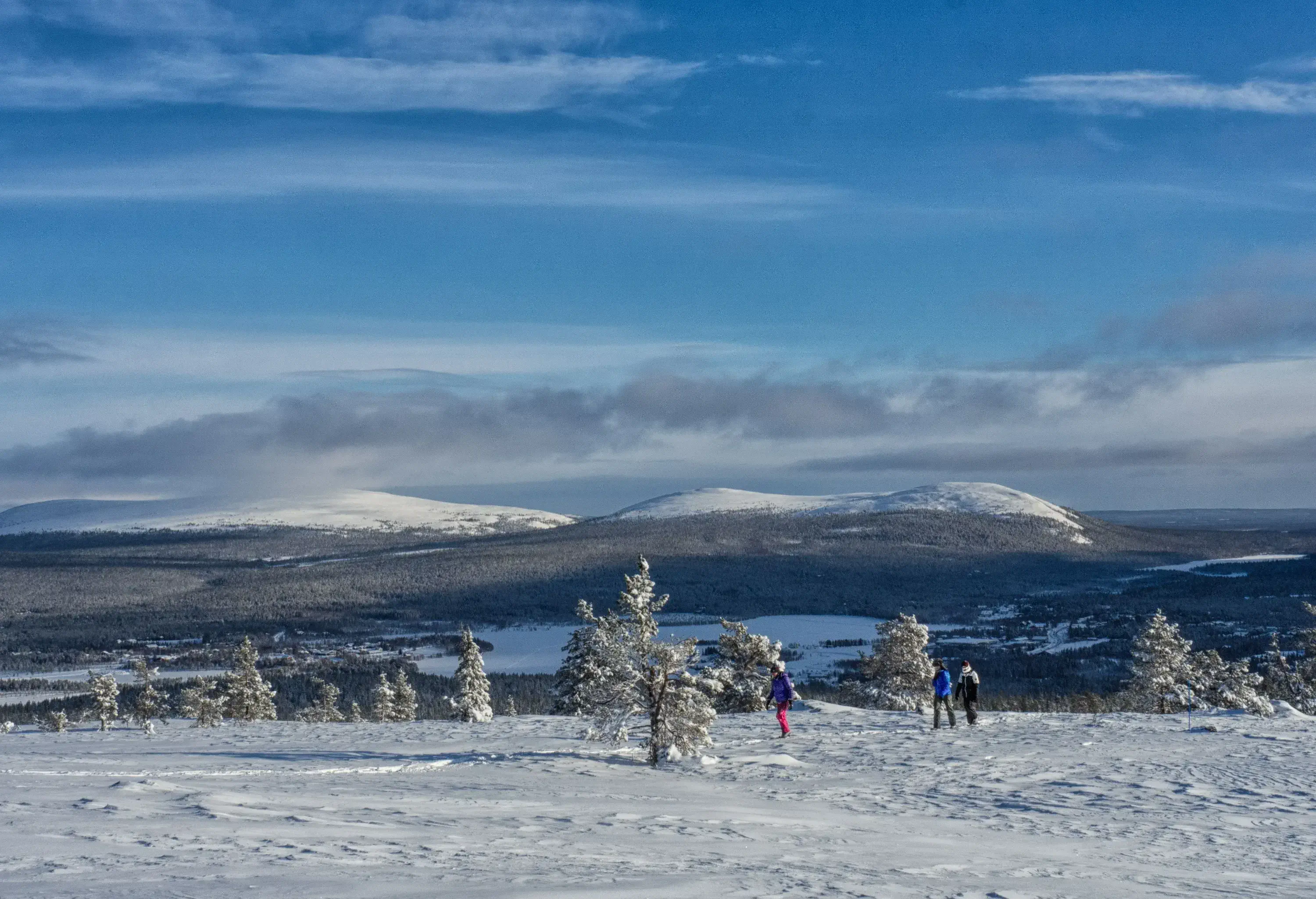 Three hikers in winter clothes traverse a snowfield with frosted trees and views of snow-capped mountains in the distance.