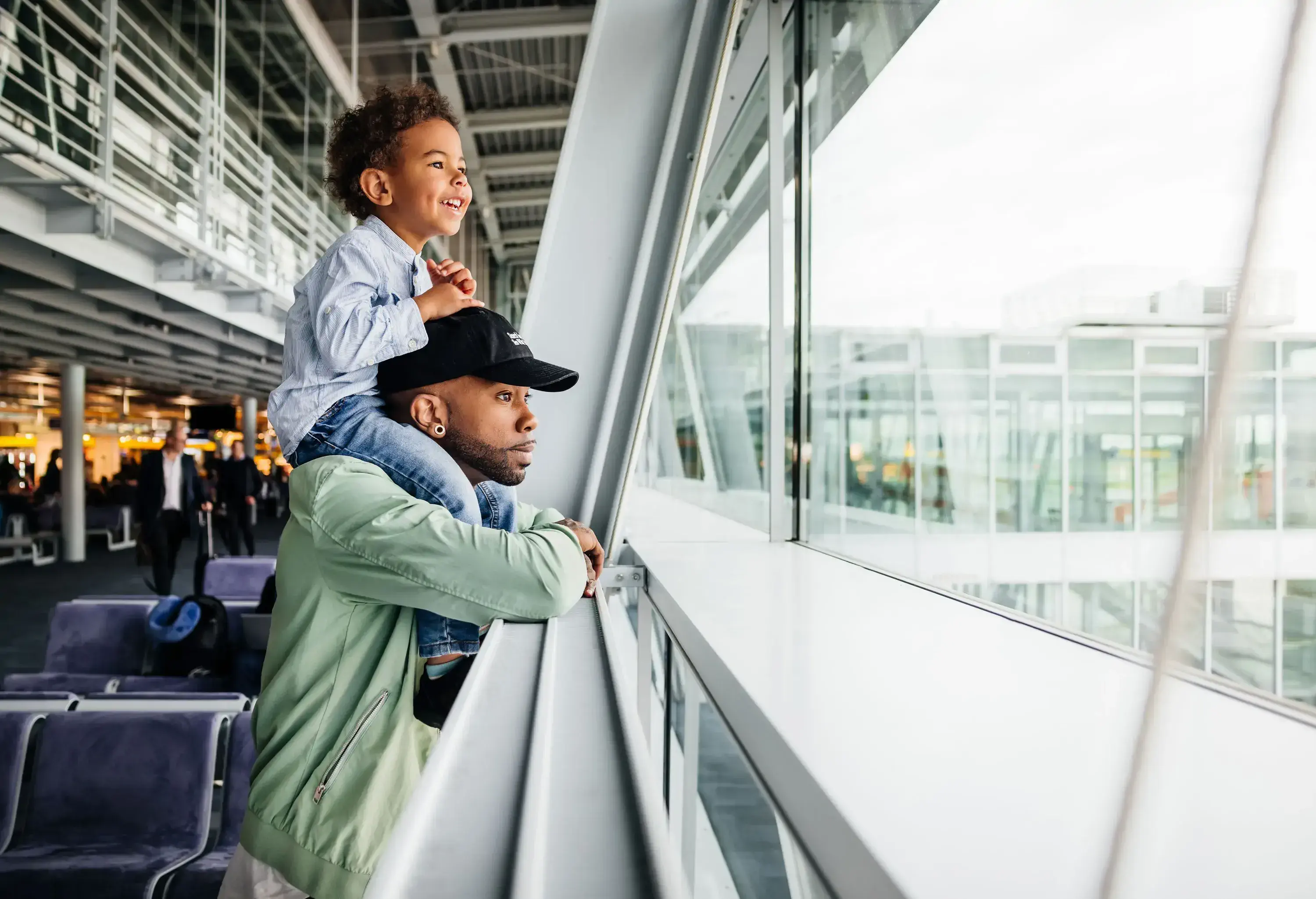A delighted child sitting on his father's shoulders at an airport, both enjoying the view from the terminal window, watching the planes, and capturing the excitement of pre-flight.
