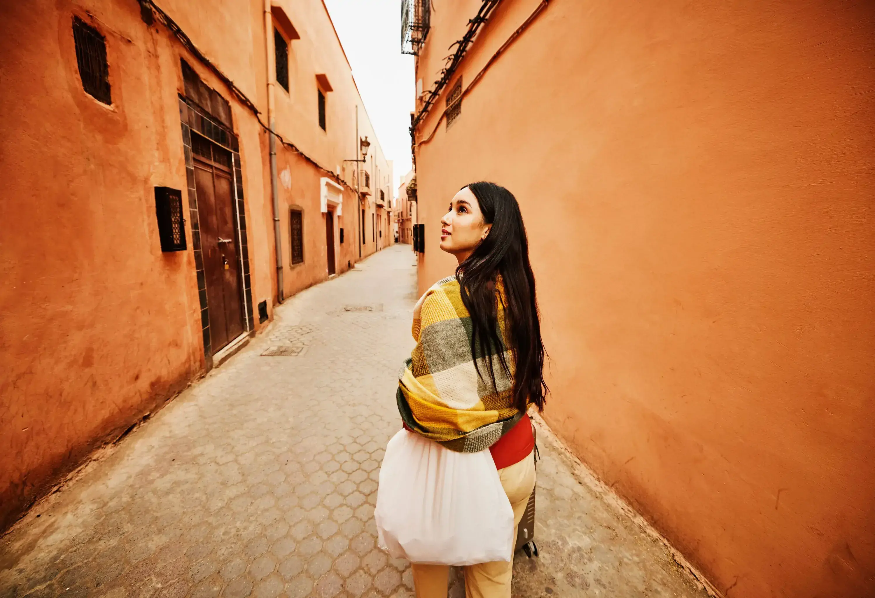 A woman looking upwards while walking through a historic-looking street with orange walls.