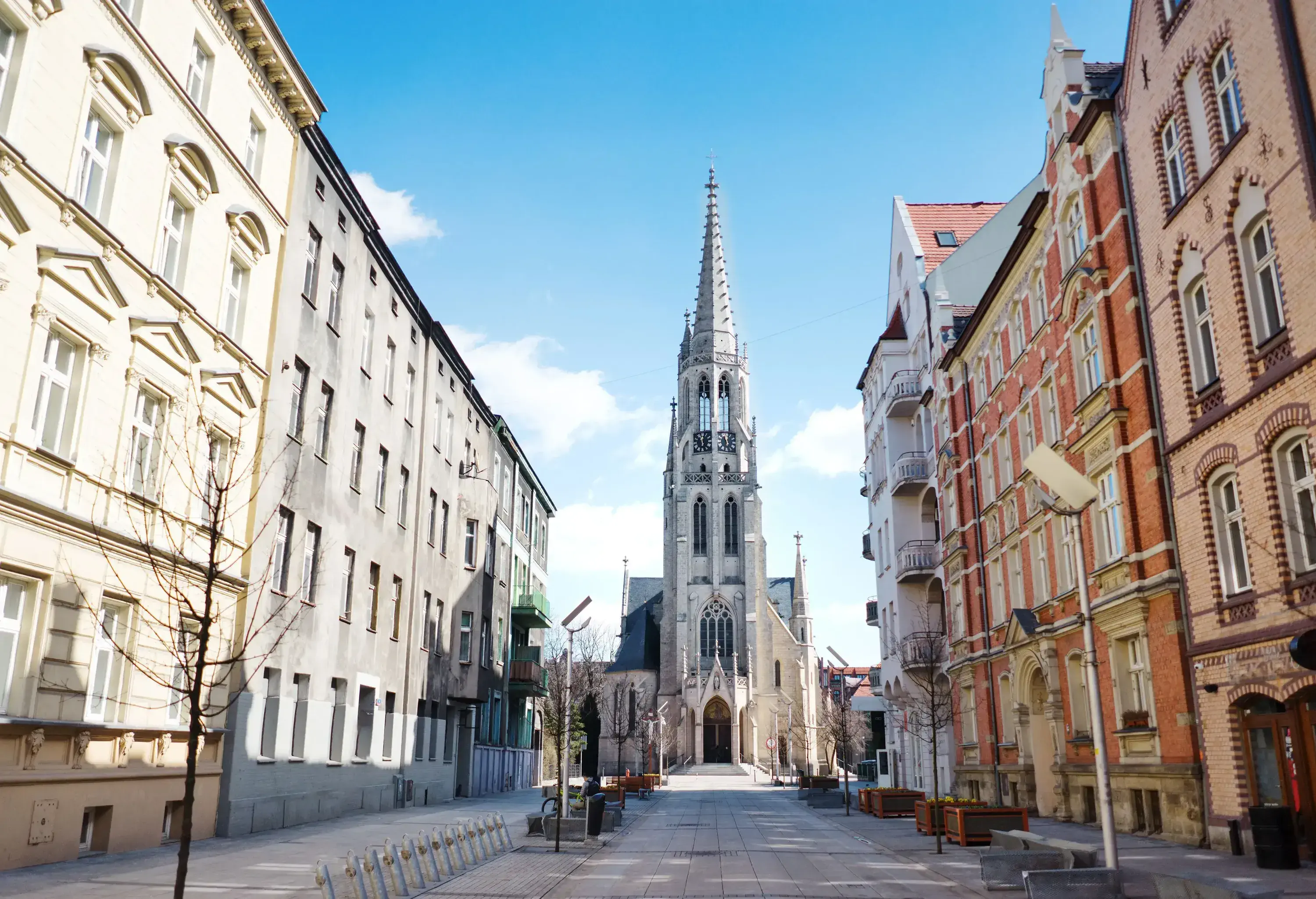 An empty street flanked by tall city buildings towards a church with a sharp spire.
