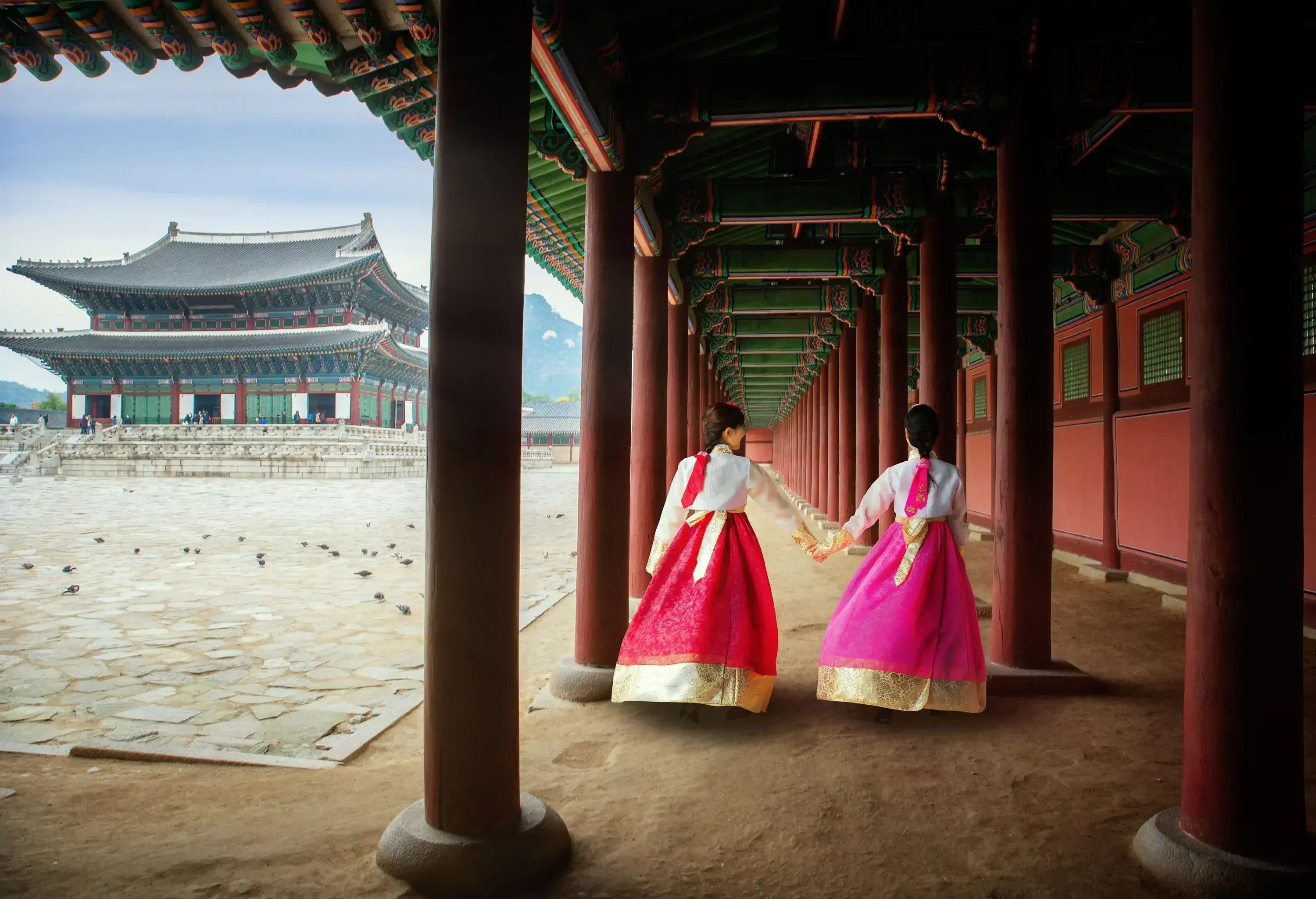 Two Korean ladies dressed in elegant Hanbok stroll along a canopied walkway adorned with vibrant red columns, as the grandeur of an ancient Korean palace provides a majestic backdrop.
