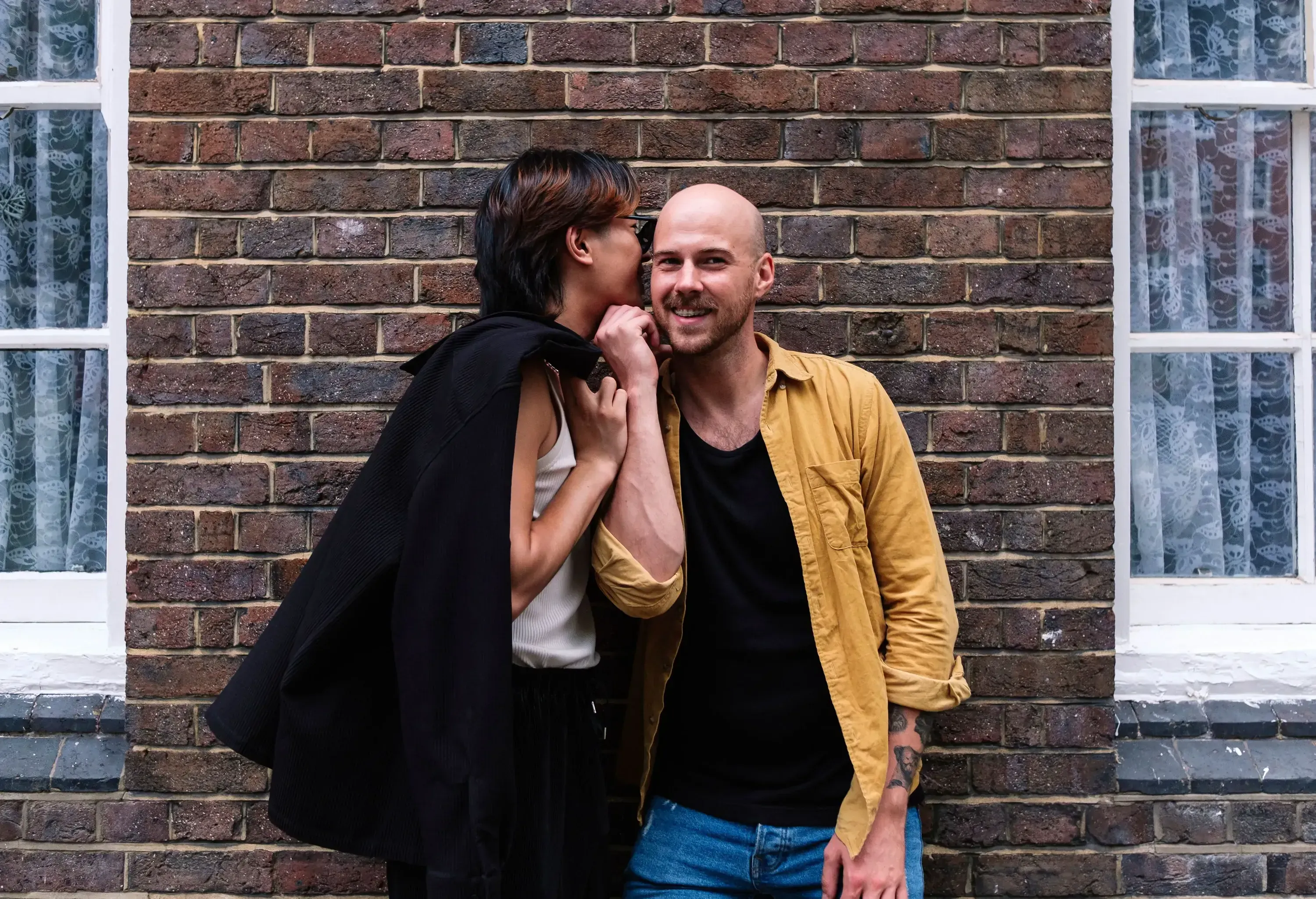 A man whispering in his smiling boyfriend's ear while leaning on a bricked wall in the centre of two windows.