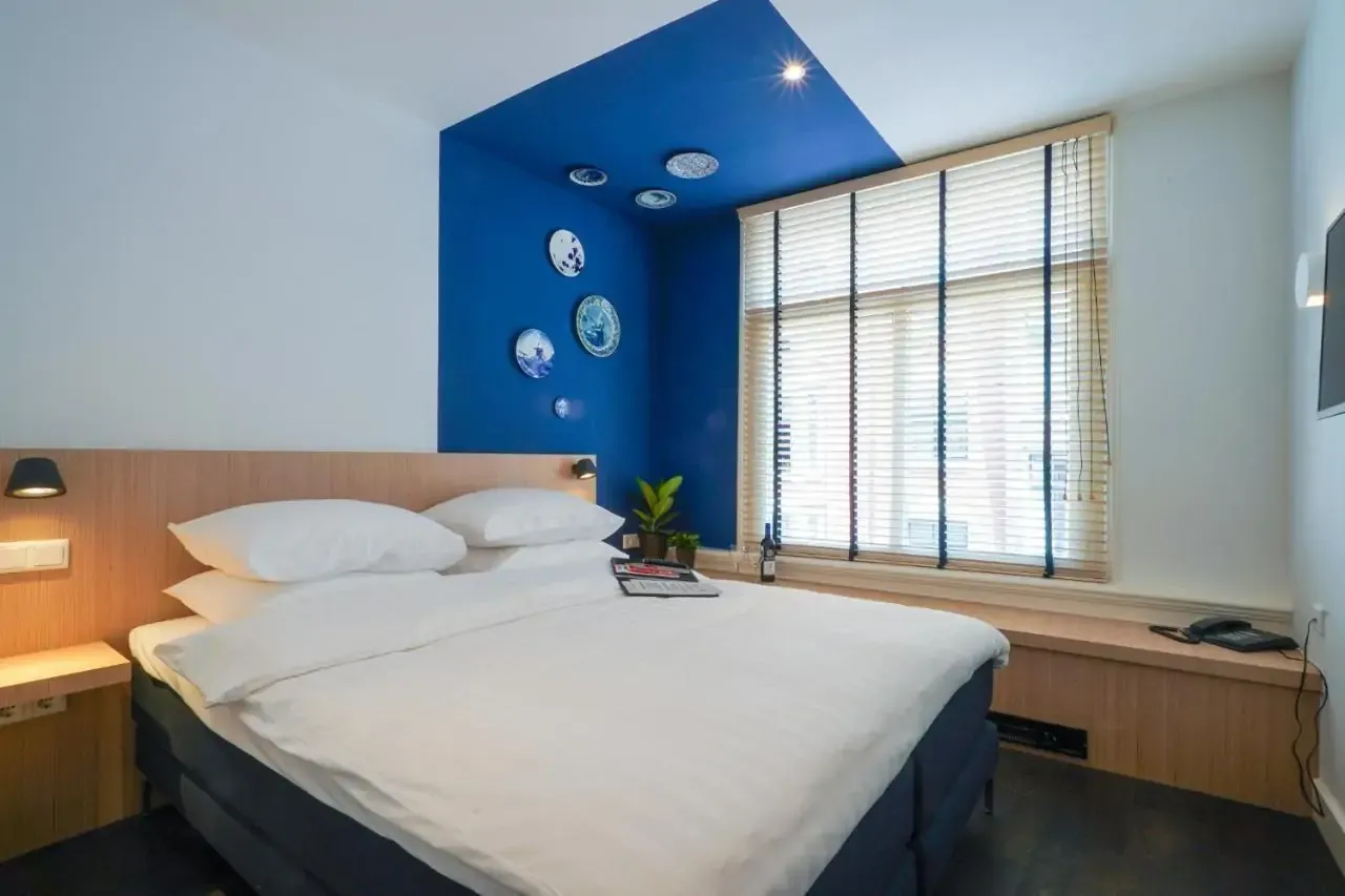 Modern hotel room with a cozy double bed featuring white linens, a striking blue accent wall with decorative plates, and a window with blinds.