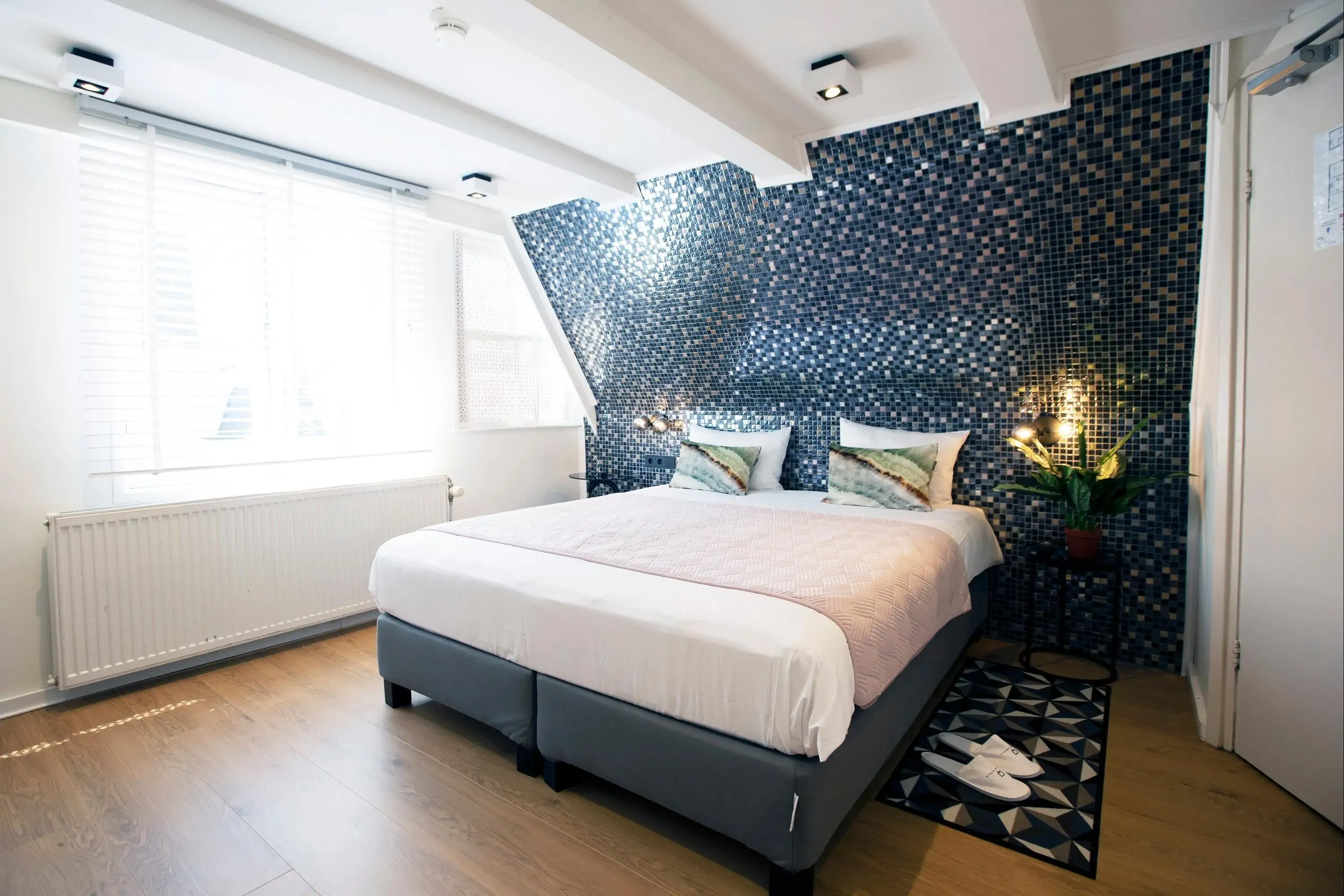 Modern bedroom with a geometric blue-tiled accent wall. A bed with pastel pillows is centered beneath, next to a leafy plant. Sunlight filters through blinds.