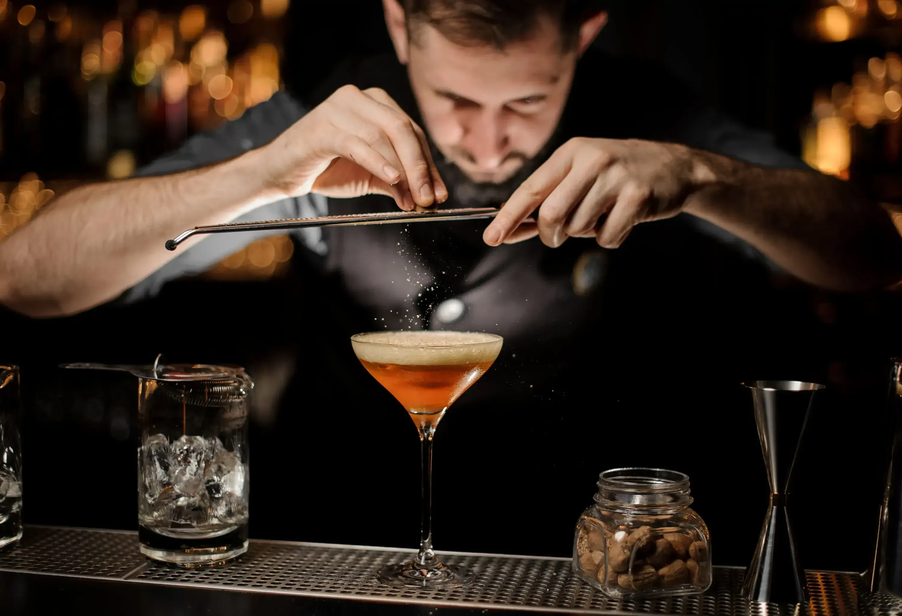 A bartender grates a nutmeg over a cocktail drink in a wine glass.