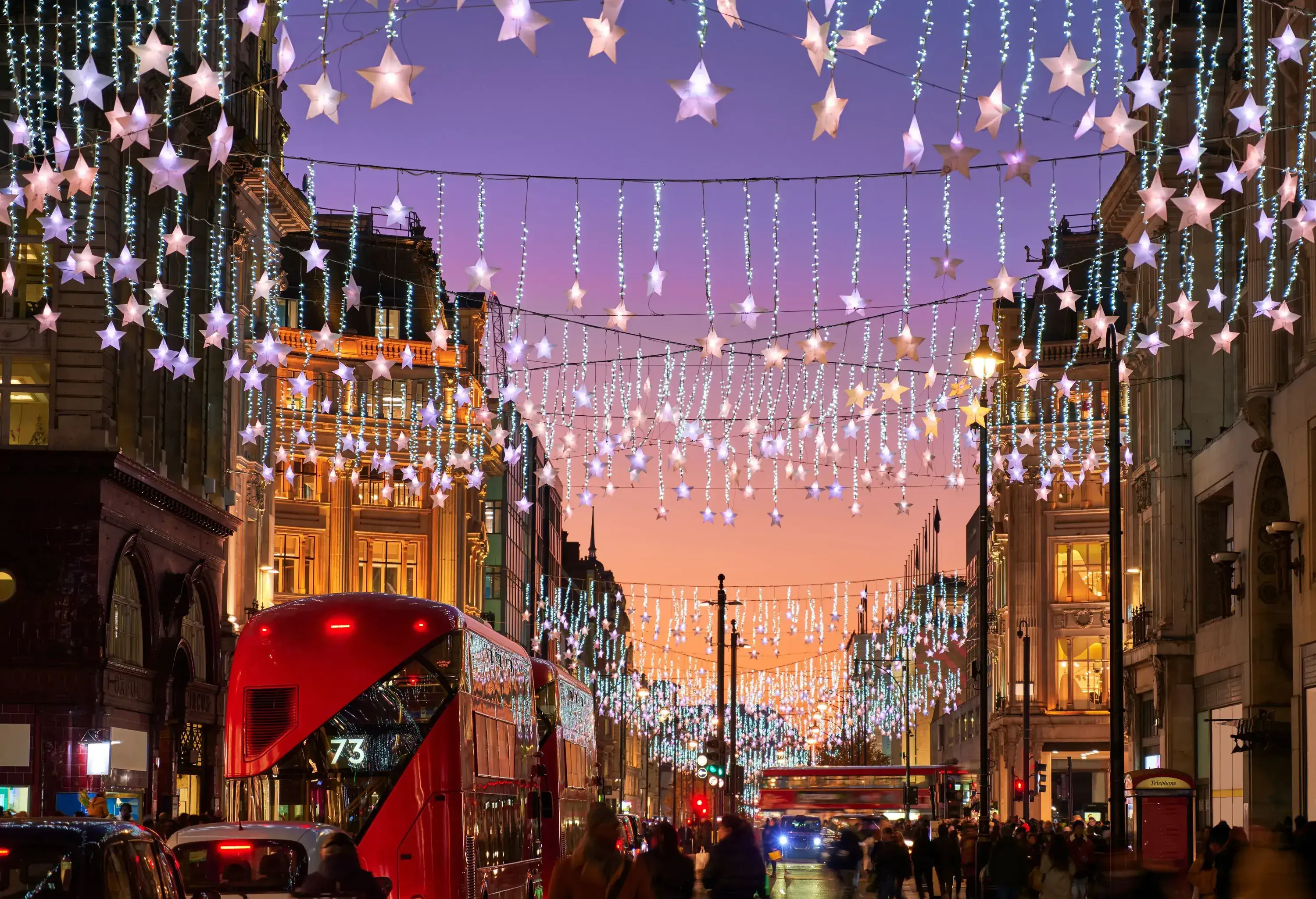 Hanging star-shaped lights illuminate a city street with buildings, a red bus, and pedestrians at dusk.