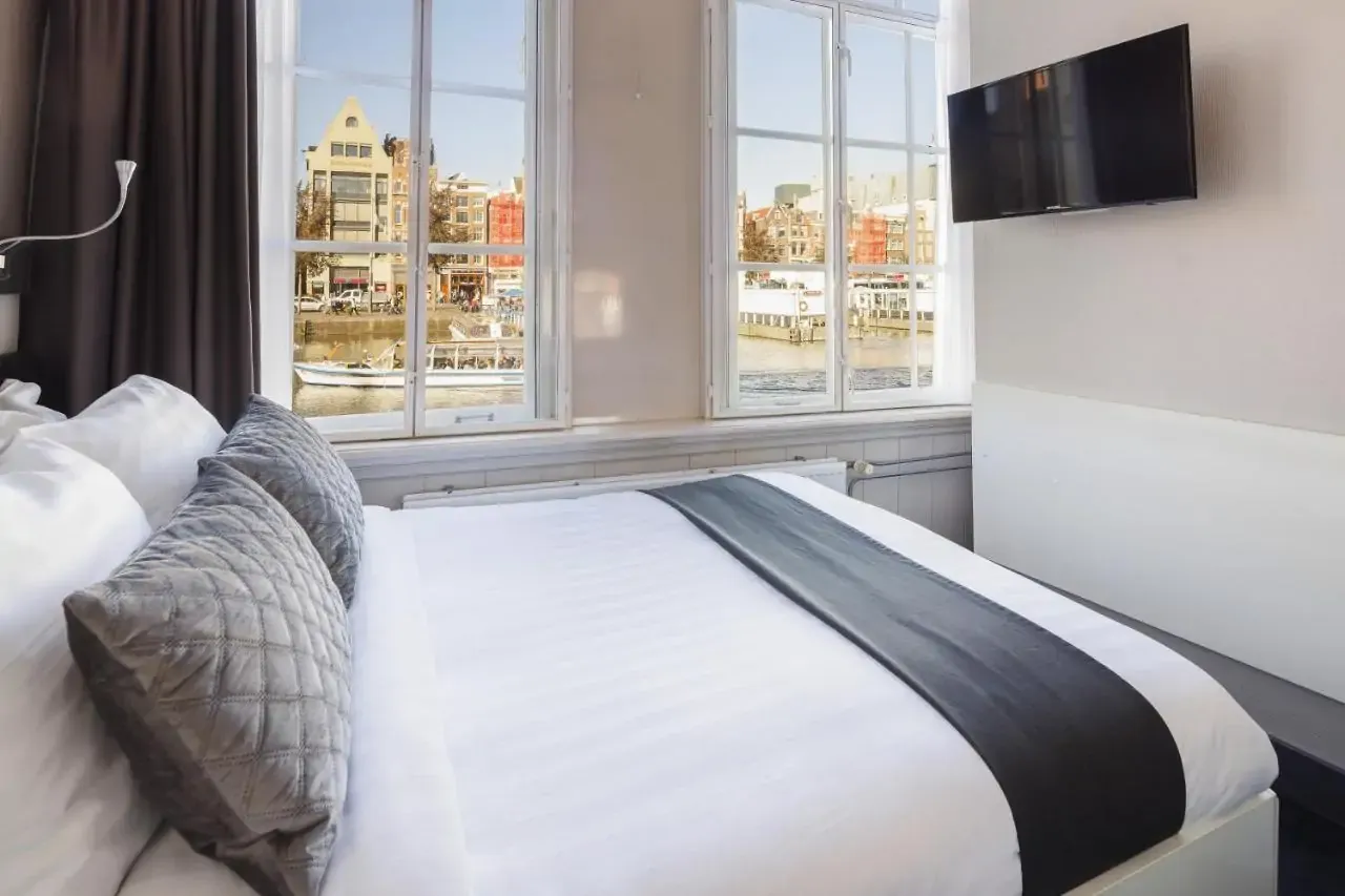 Modern hotel room with a neatly made bed, gray accents, and a wall-mounted TV. Large windows reveal a scenic canal view with charming buildings.