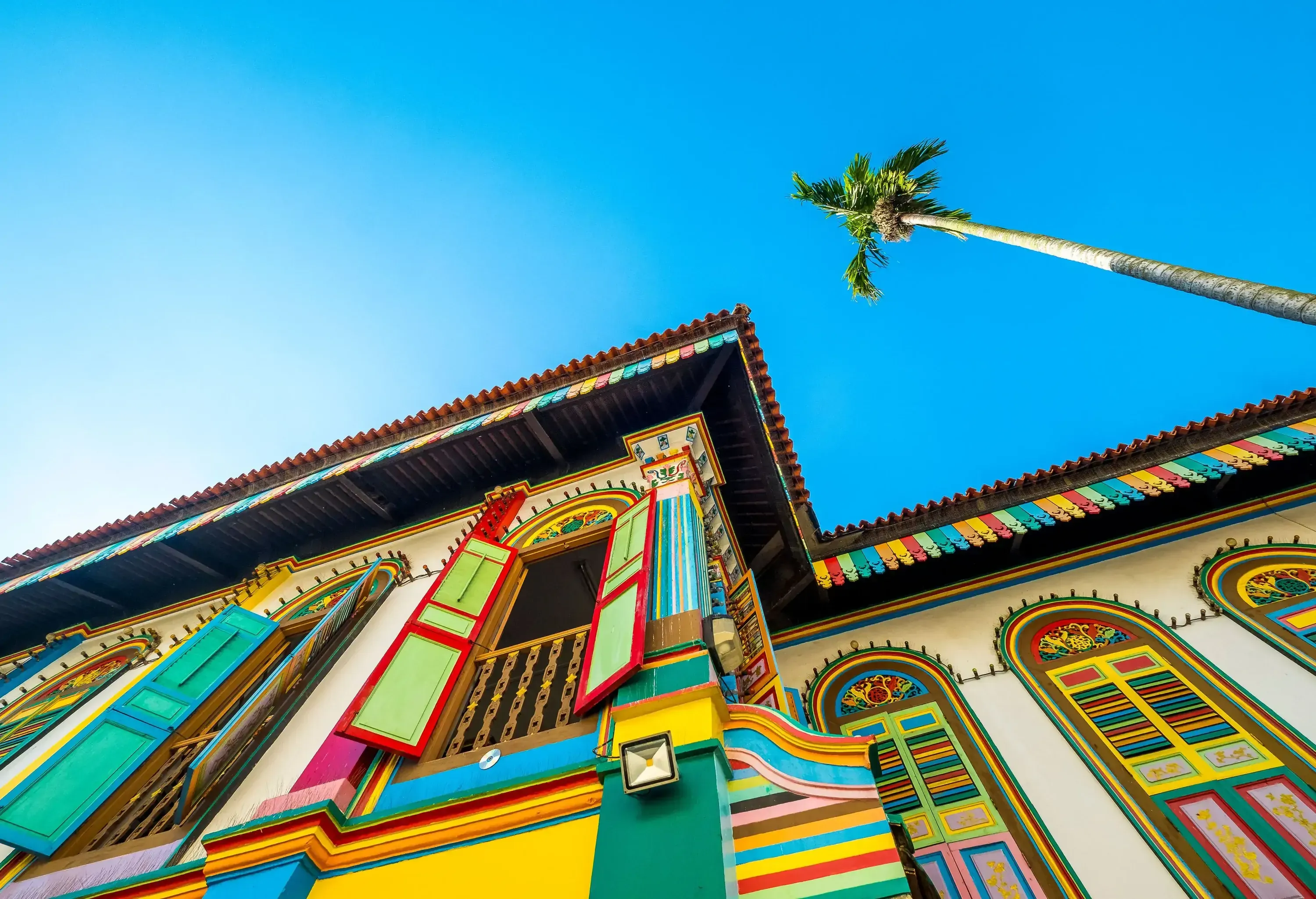 A vibrant, traditional building with colorful facade and windows, viewed from a low angle against a clear blue sky and a palm tree.