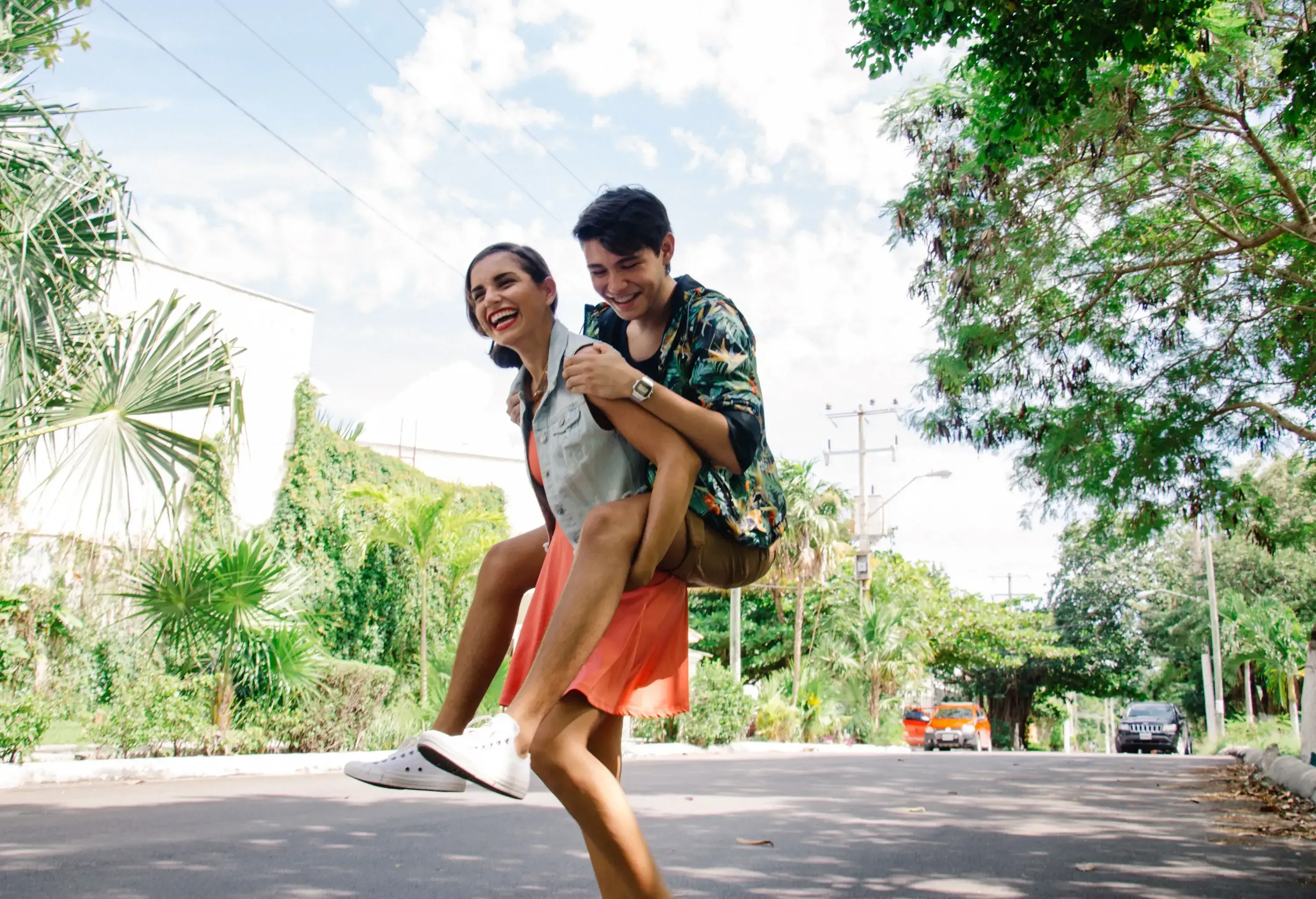 Amidst a charming street lined with trees and plants, a couple shares a moment of joy and playfulness as one gives the other a piggyback ride.